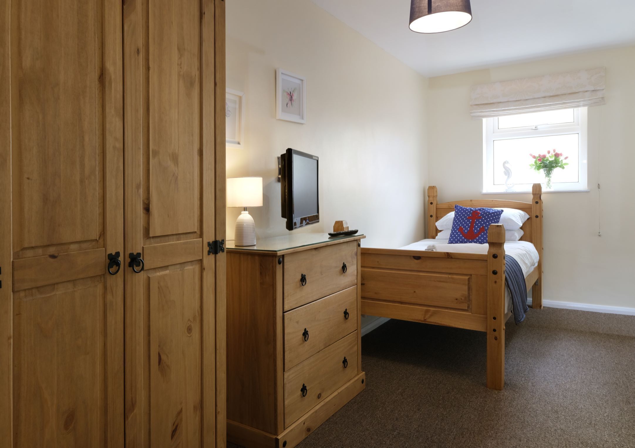 A bedroom with a wooden wardrobe, a bedside table with a lamp, a bed with a blue pillow, and a window with a view of flowers.