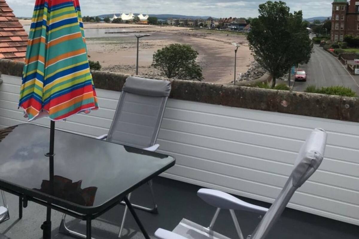 A balcony with a striped umbrella, two white chairs, and a table.