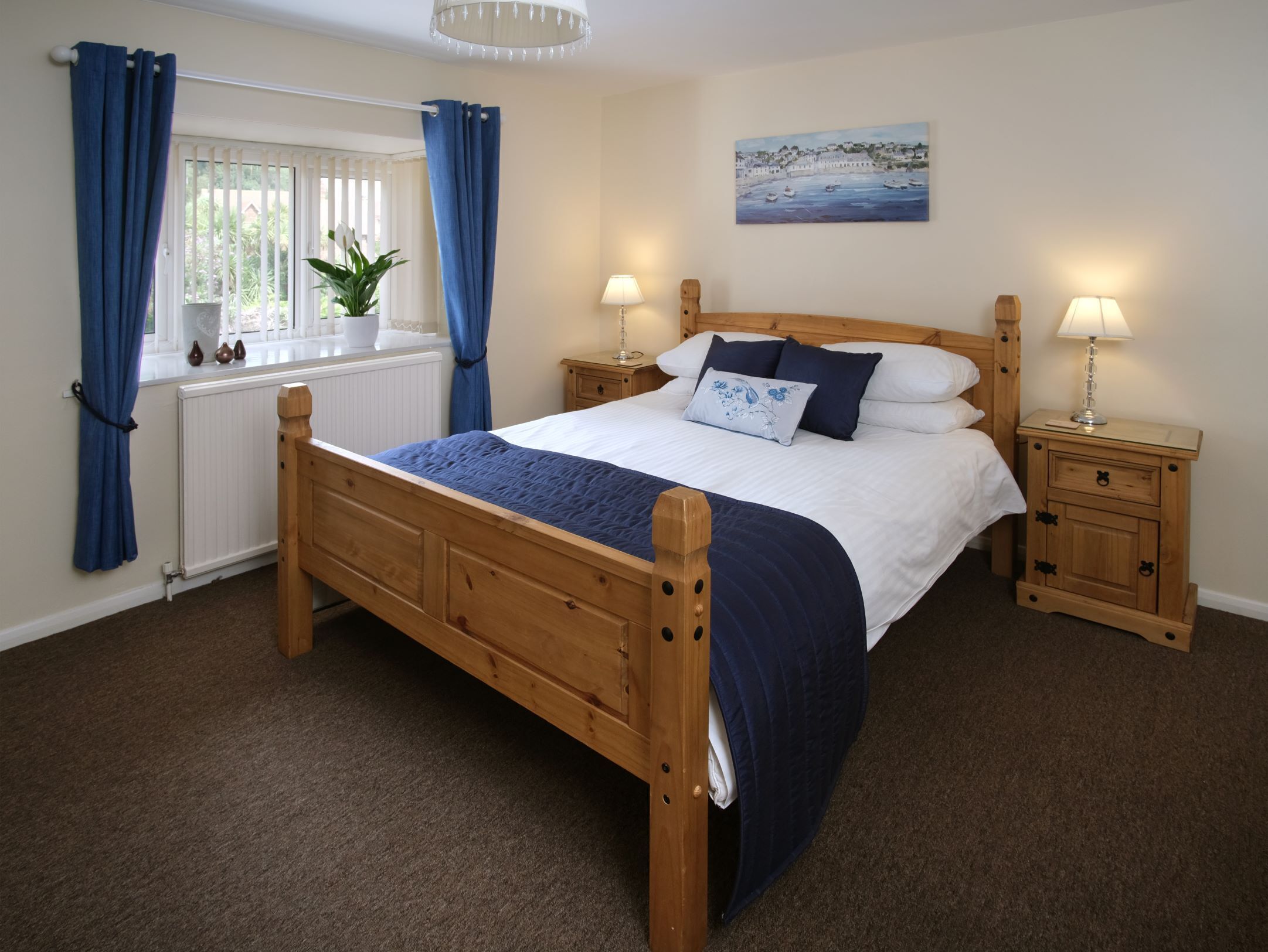 A bedroom with a wooden bed, two bedside tables with lamps, a window with curtains, a radiator, and a painting on the wall.