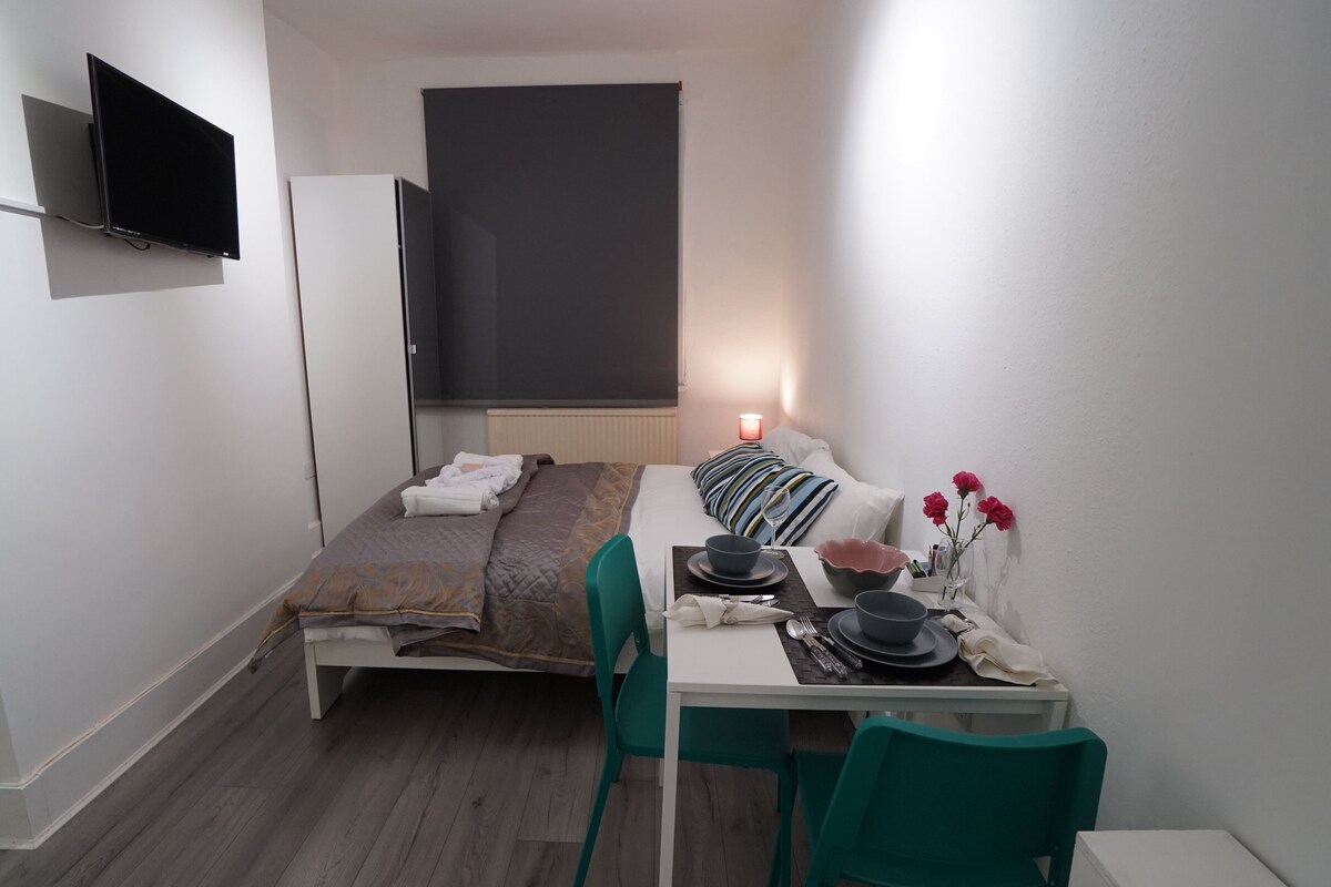 Habitación de hotel compacta con cama, una mesa de comedor para dos personas, un televisor de pantalla plana en la pared y una mesita con un jarrón de flores.