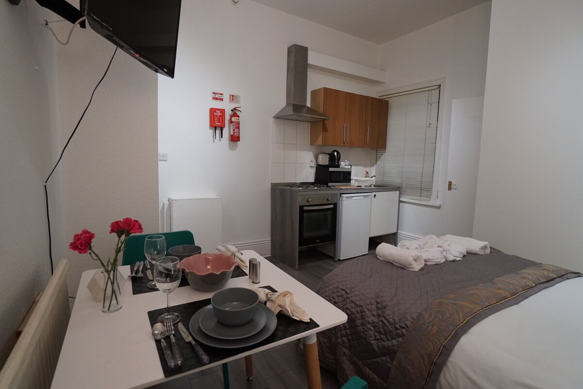 Habitación de hotel compacta con kitchenette, zona de comedor con una mesa para dos personas y cama con una colcha grande.
