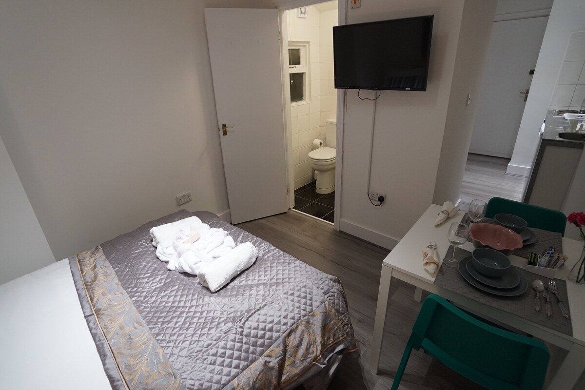 Una habitación de hotel con cama, televisor montado en la pared, zona de comedor con mesa y sillas, y baño visible a través de una puerta abierta.