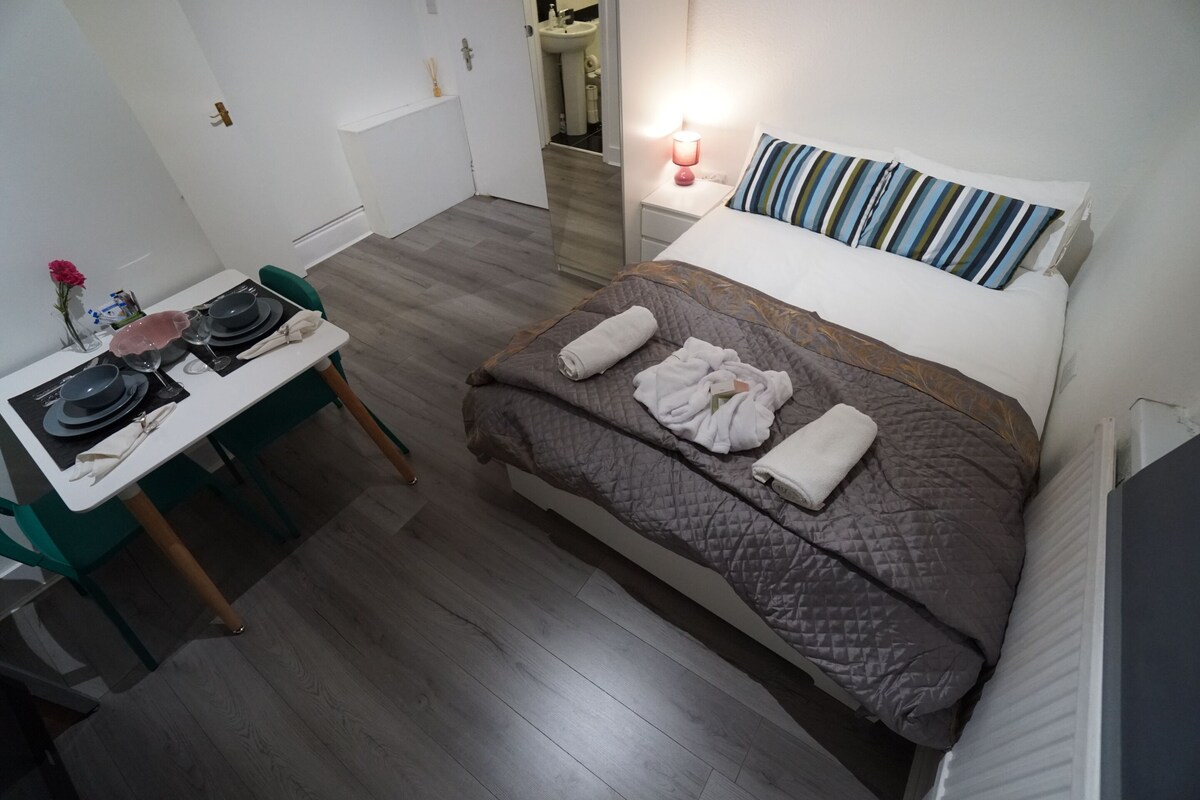 Una habitación pequeña con cama, mesita de noche y una silla junto a una pequeña mesa.