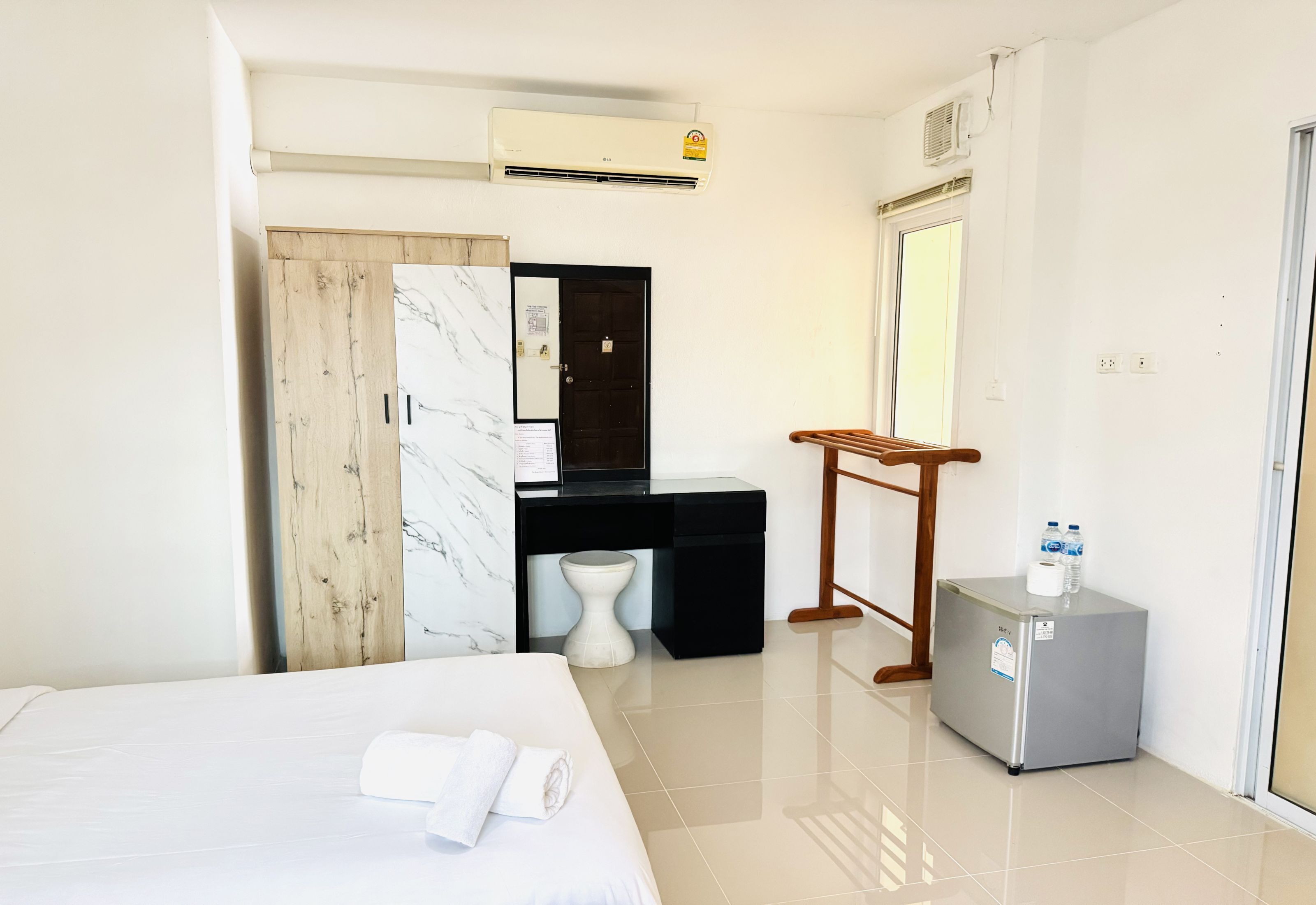A hotel room with a bed, a desk with a chair, a small refrigerator, a toilet, and an air conditioning unit.