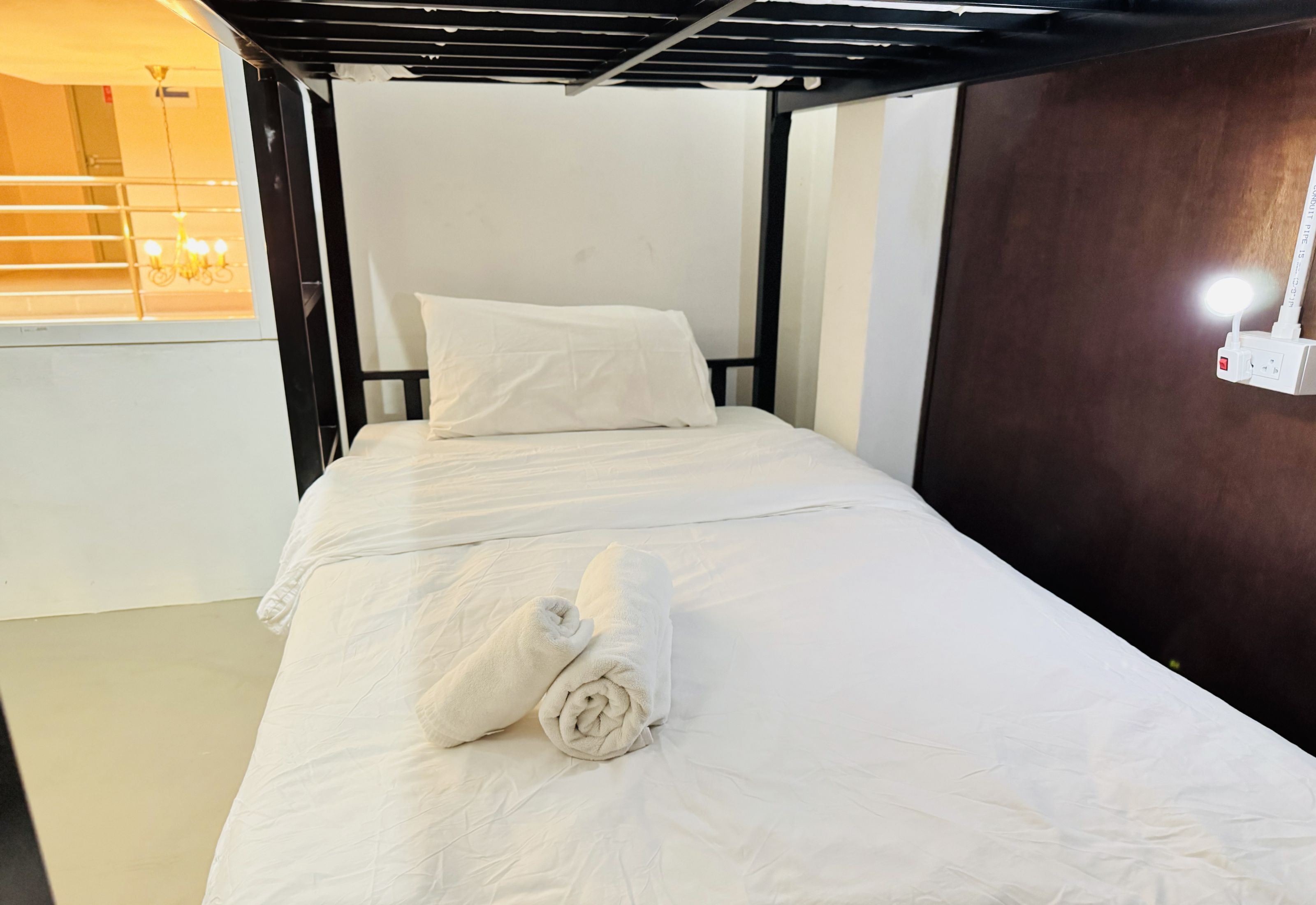 A single bed with white bedding and a rolled towel, a wooden headboard, and a wall-mounted light fixture.