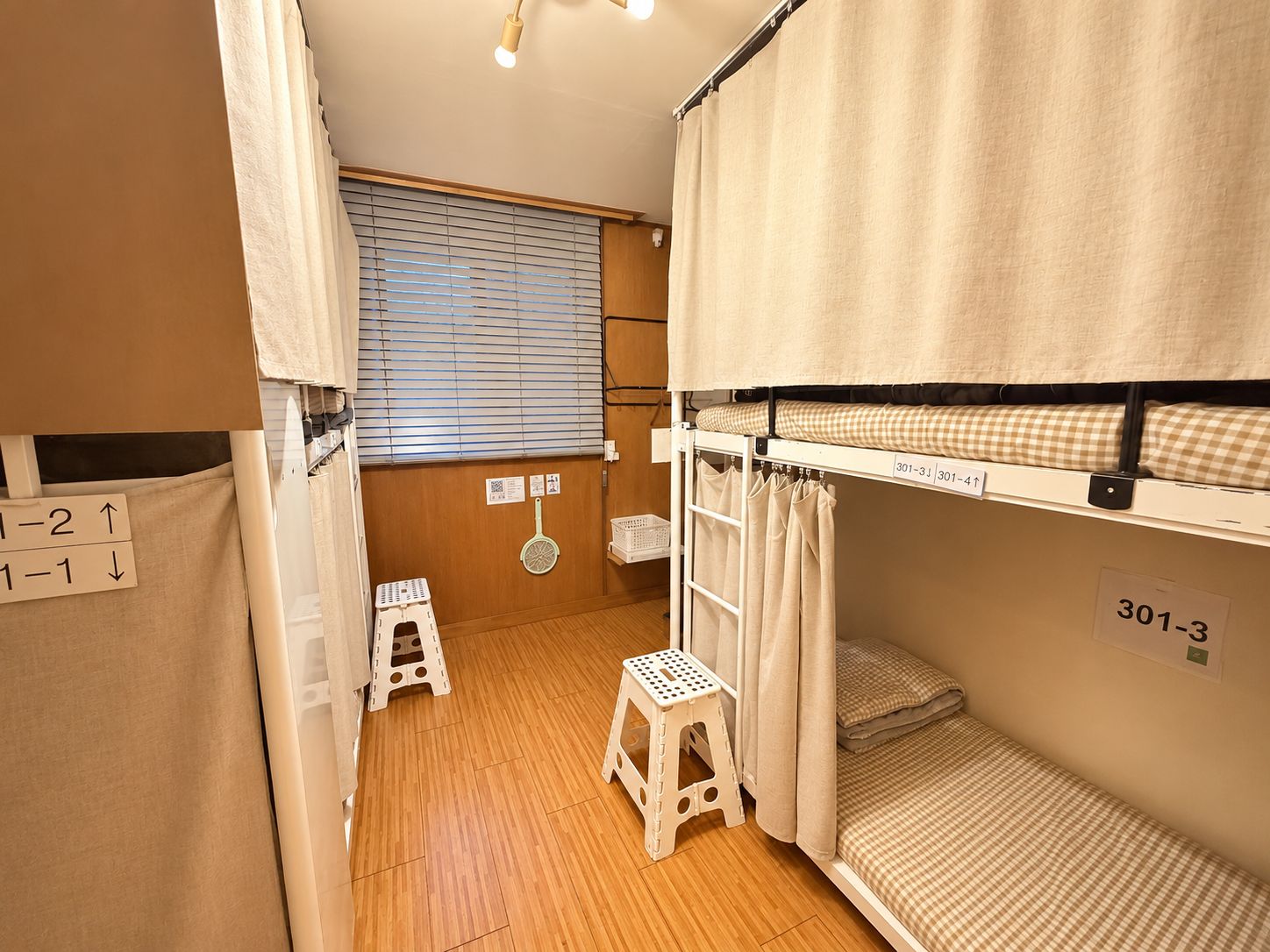 A dormitory room with bunk beds, a wooden floor, a window with blinds, a small stool, and a wall-mounted shelf.