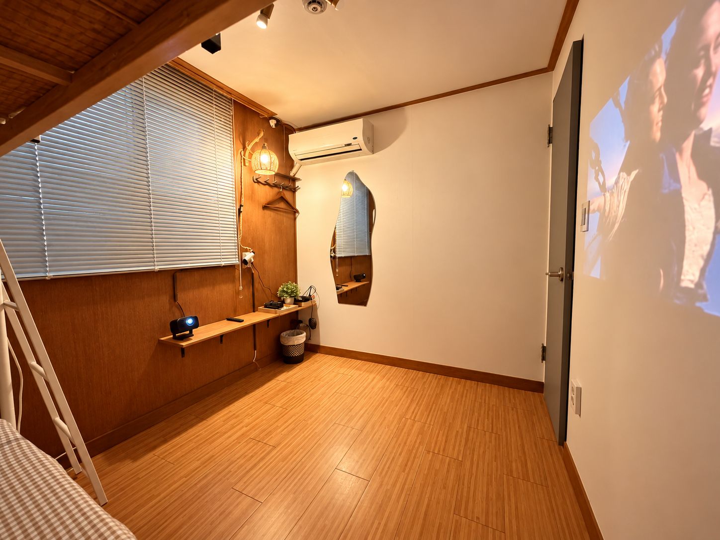 Una habitación con piso de madera, una cama, un escritorio, una silla, un ventanal con persianas, un equipo de aire acondicionado y un proyector montado en la pared.