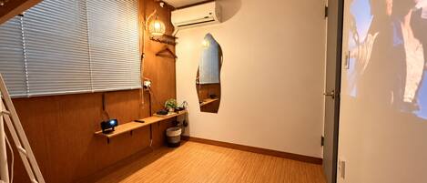 Una habitación con piso de madera, una cama, un escritorio, una silla, un ventanal con persianas, un equipo de aire acondicionado y un proyector montado en la pared.