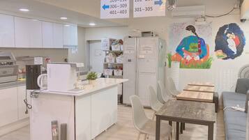 A modern kitchen with white cabinets, a coffee machine, and a refrigerator. There are signs hanging from the ceiling indicating room numbers.
