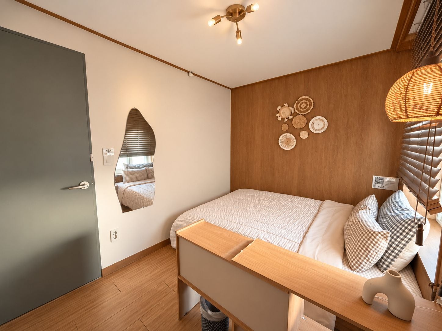 A modern bedroom with a wooden bed, a mirror, a wall-mounted light fixture, and a decorative wall piece.