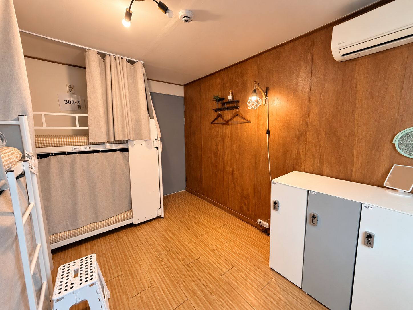 A compact room with a bunk bed, a small kitchenette, a wooden wall, and a ceiling-mounted air conditioner.