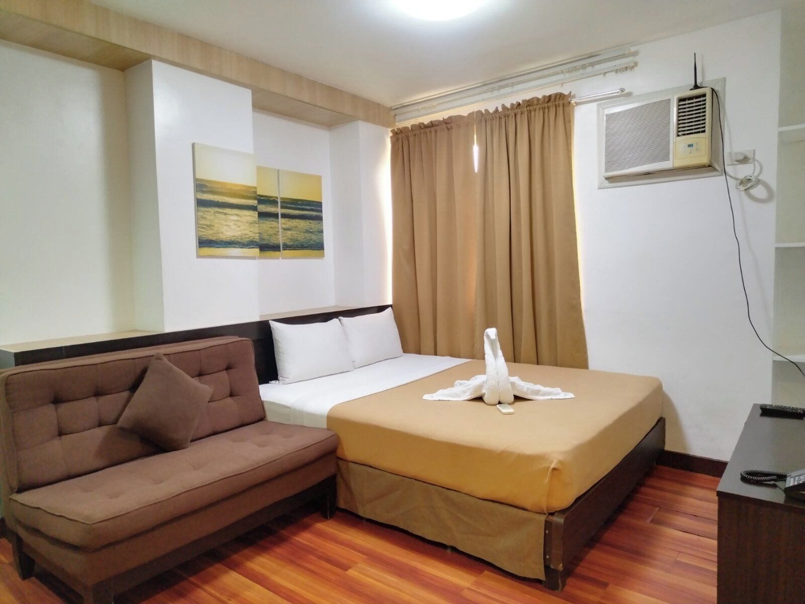 A hotel room with a bed, a brown sofa, a wooden floor, a painting on the wall, and an air conditioning unit.