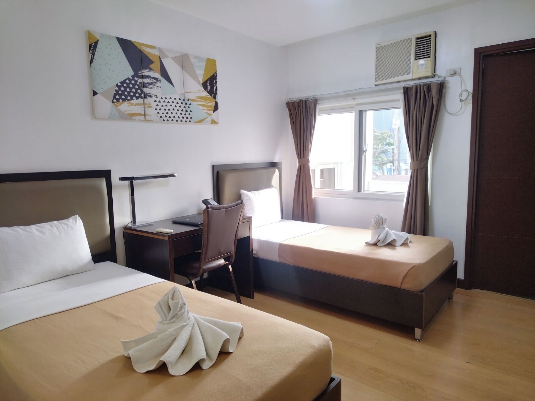 A hotel room with two beds, a desk, a chair, a window with curtains, an air conditioning unit, and a wall art piece.