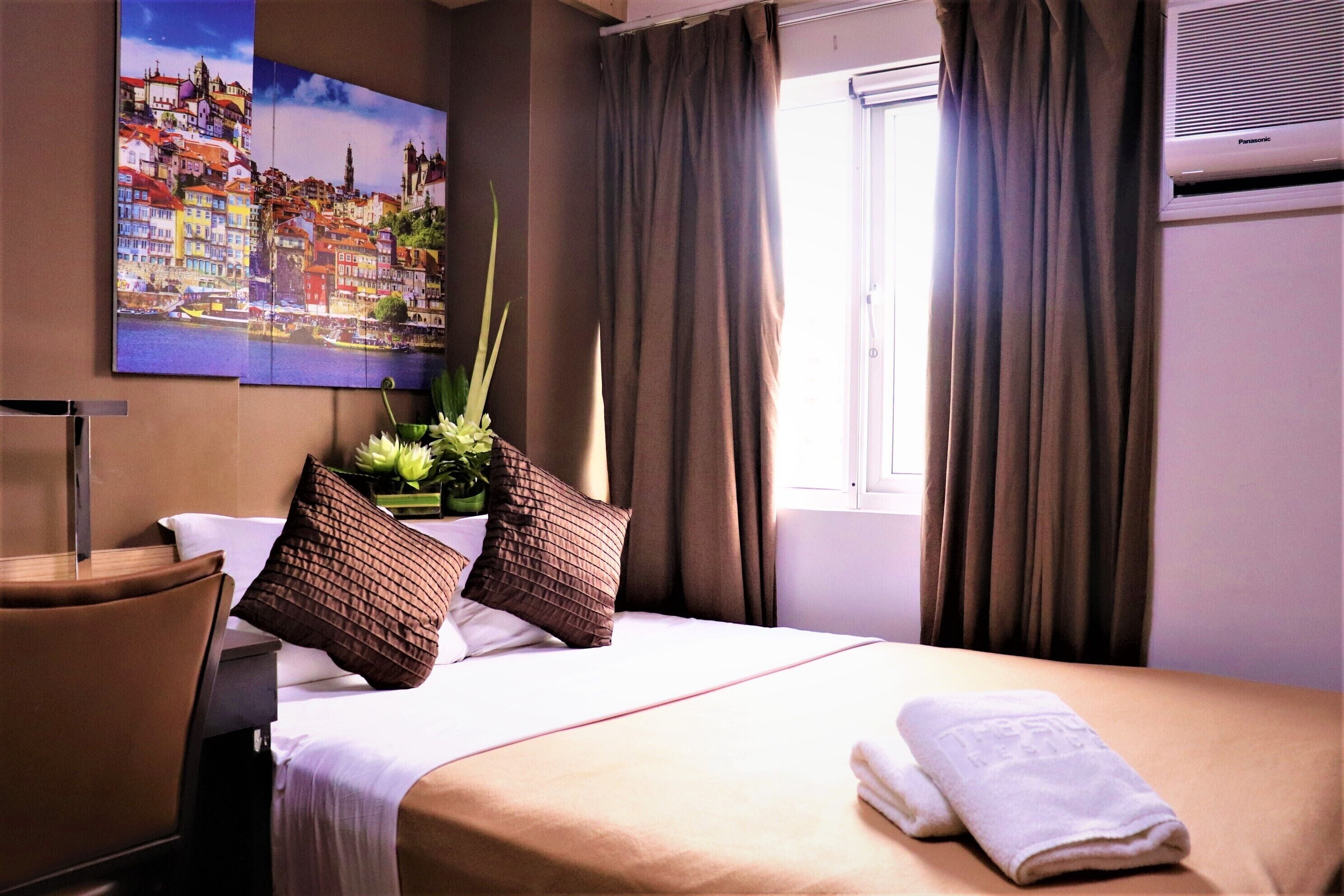 Una habitación de hotel con cama, ventana con cortinas, aire acondicionado de pared y un cuadro de un paisaje urbano.