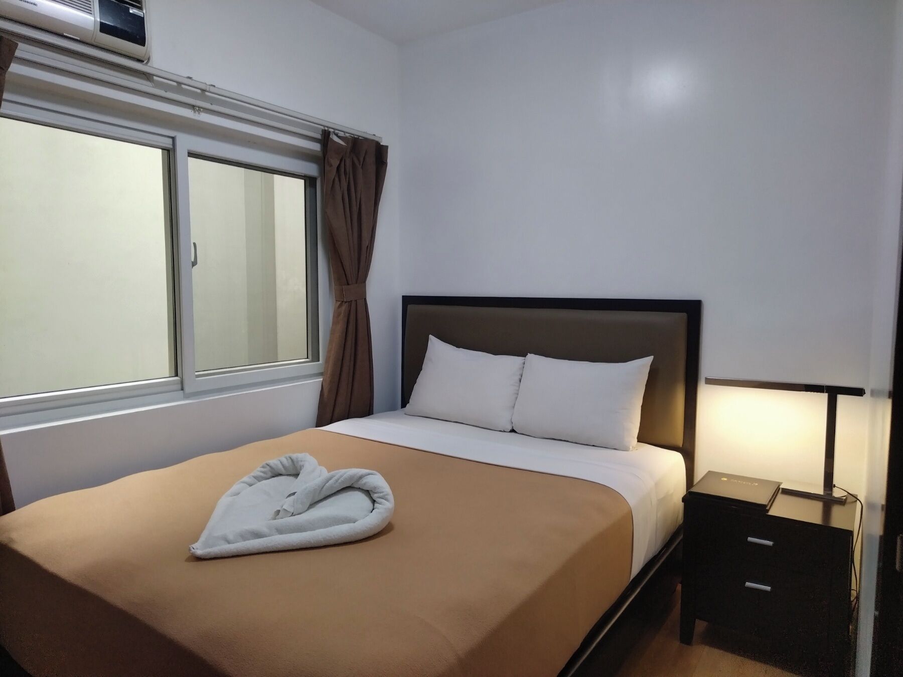 A hotel room with a bed, a window with curtains, a bedside table with a lamp, and a wall-mounted air conditioner.