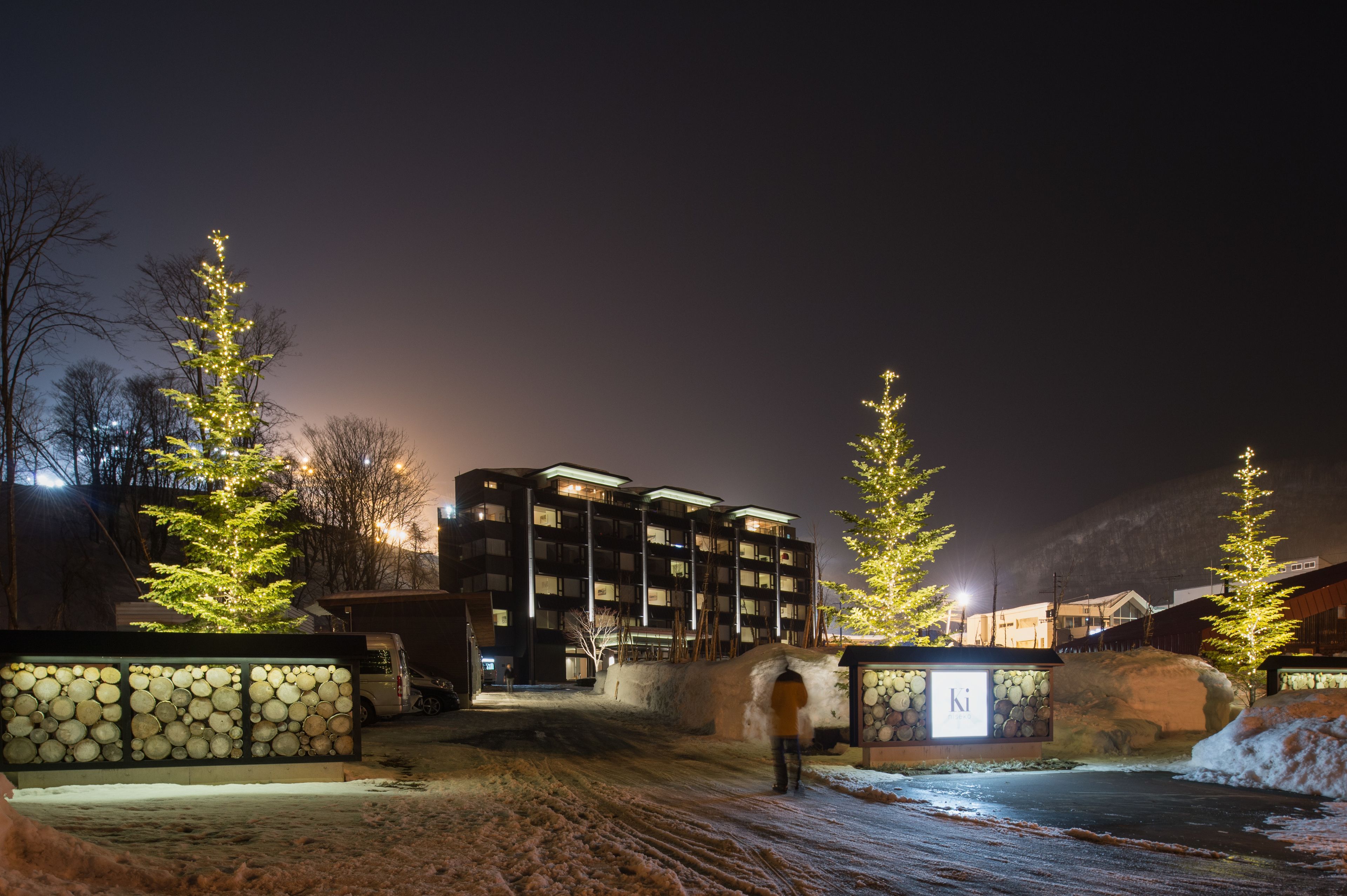 Un edificio de varios pisos, suelo cubierto de nieve y árboles de Navidad con luces.