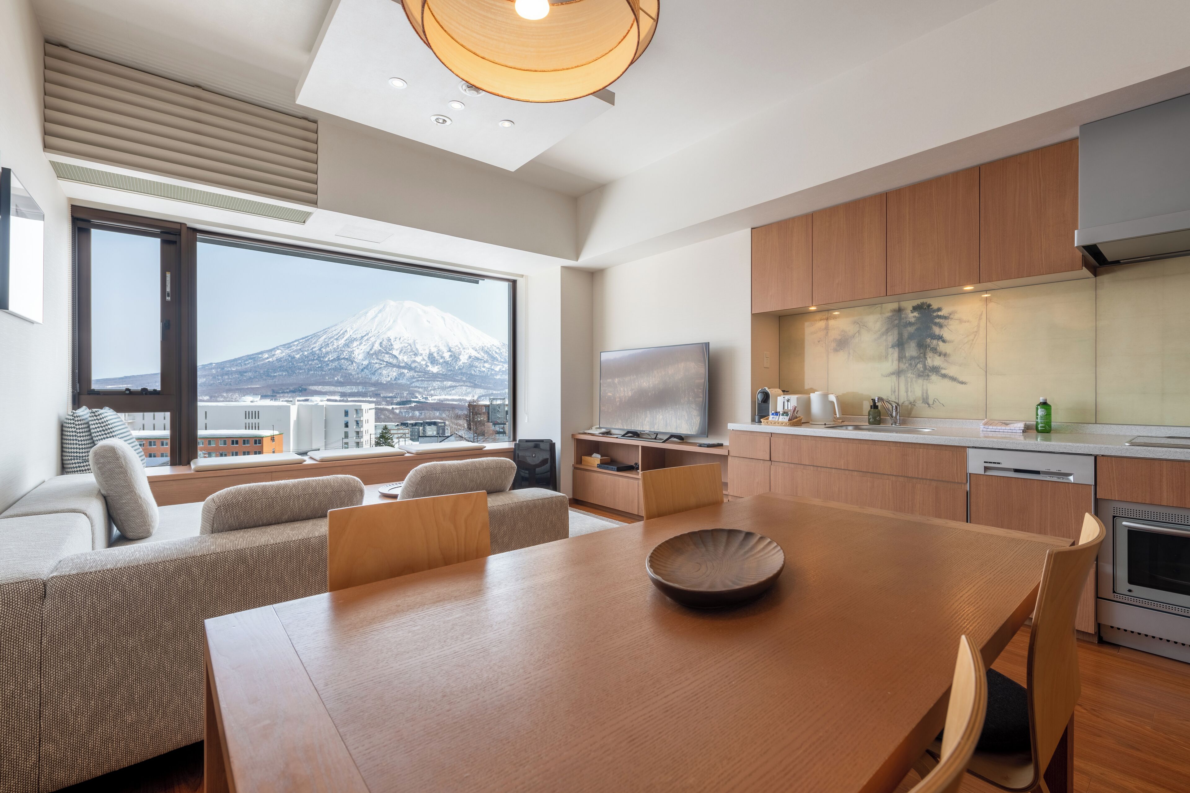 A modern kitchen with wooden cabinets, a dining area with a wooden table, and a large window offering a view of a mountain.