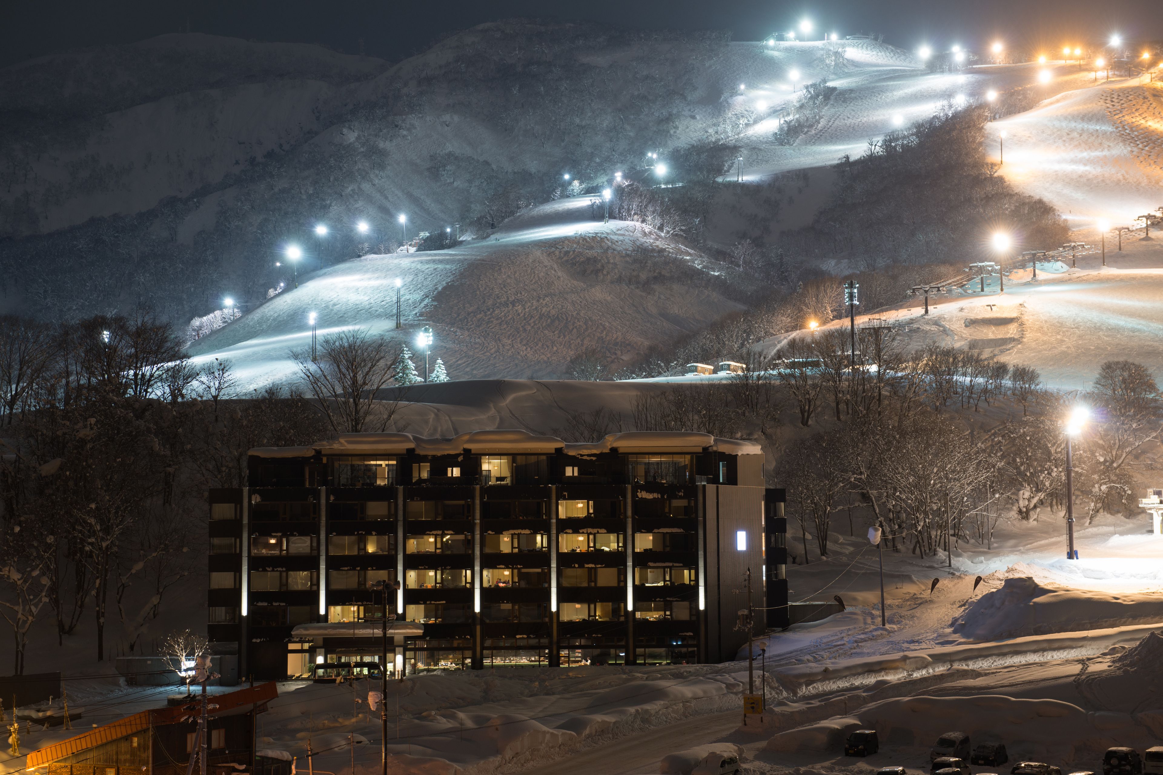 Un hôtel à plusieurs étages, dont les fenêtres sont illuminées, situé dans un paysage enneigé avec une piste de ski en arrière-plan.