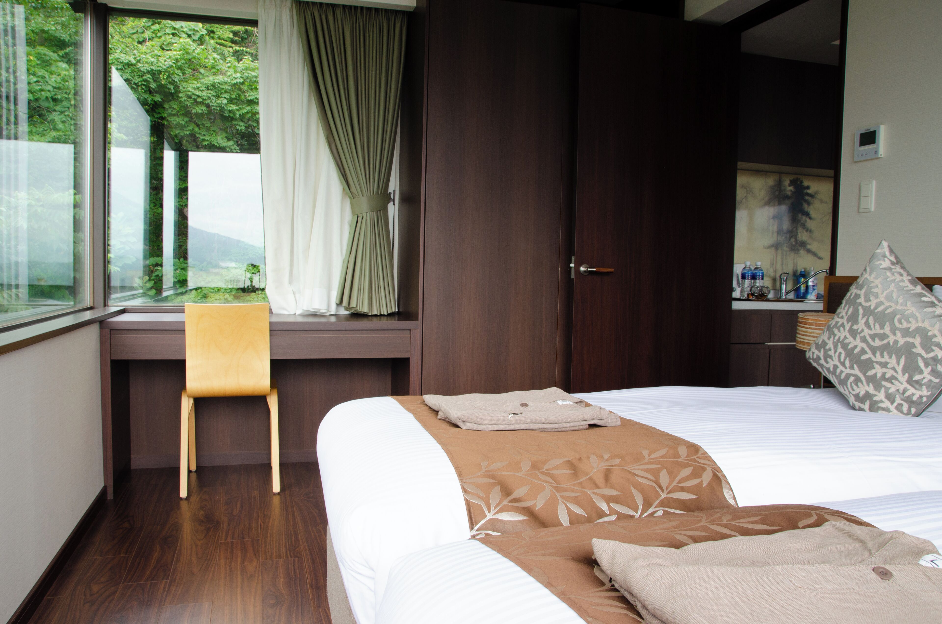 Una camera d'hotel con un letto, una scrivania, una sedia, una finestra con tende e vista sul verde.