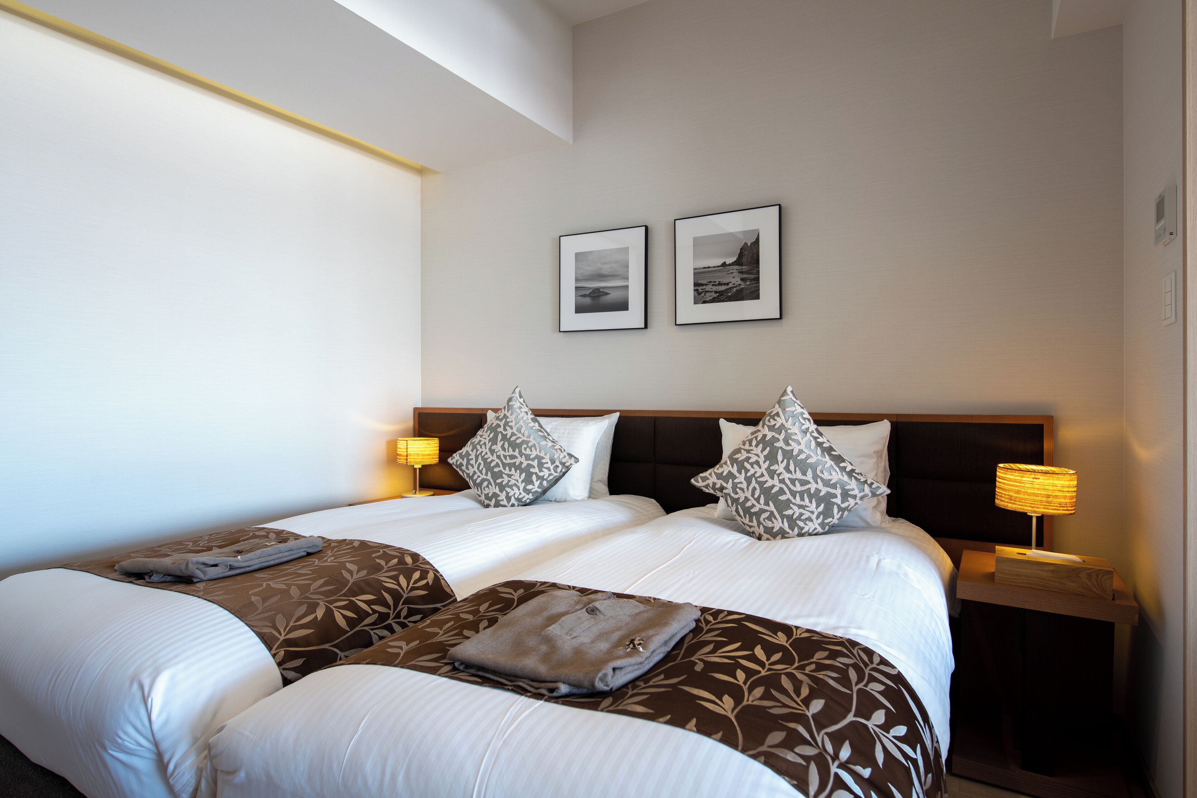 A hotel room with two beds, a headboard, two bedside lamps, and two framed pictures on the wall.