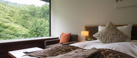 Una habitación de hotel con cama, mesita de noche, lámpara y ventana con vistas a un bosque.