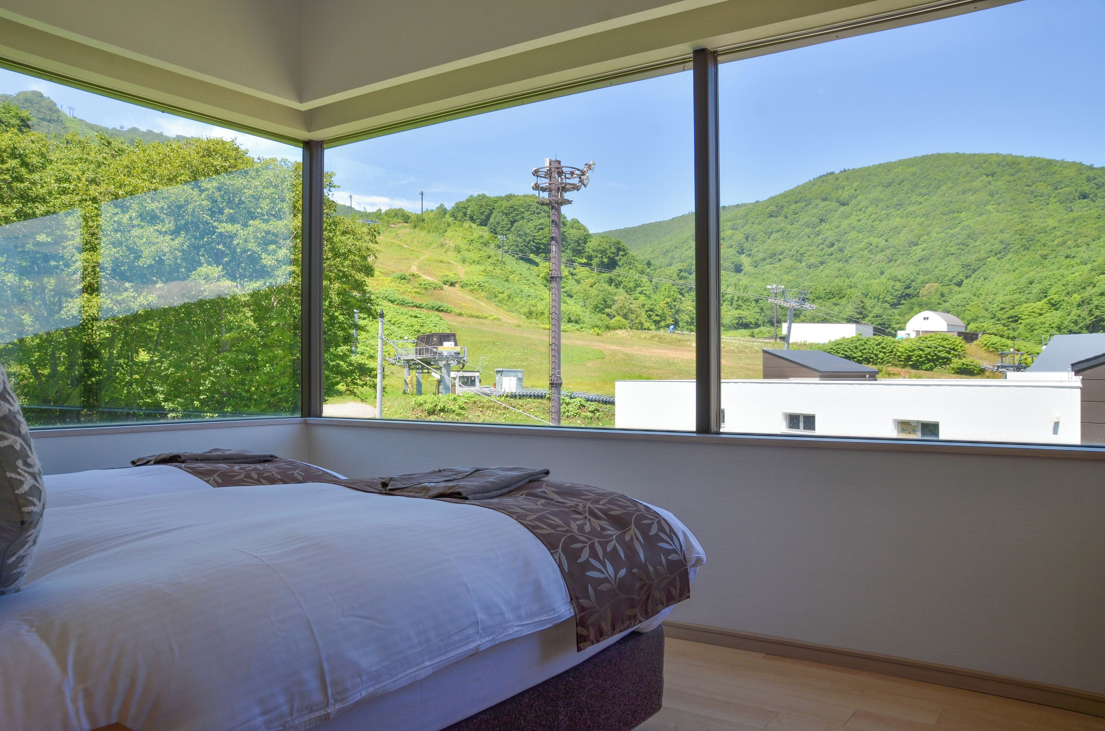 A bedroom with a large window offering a view of a green hillside and a white building.