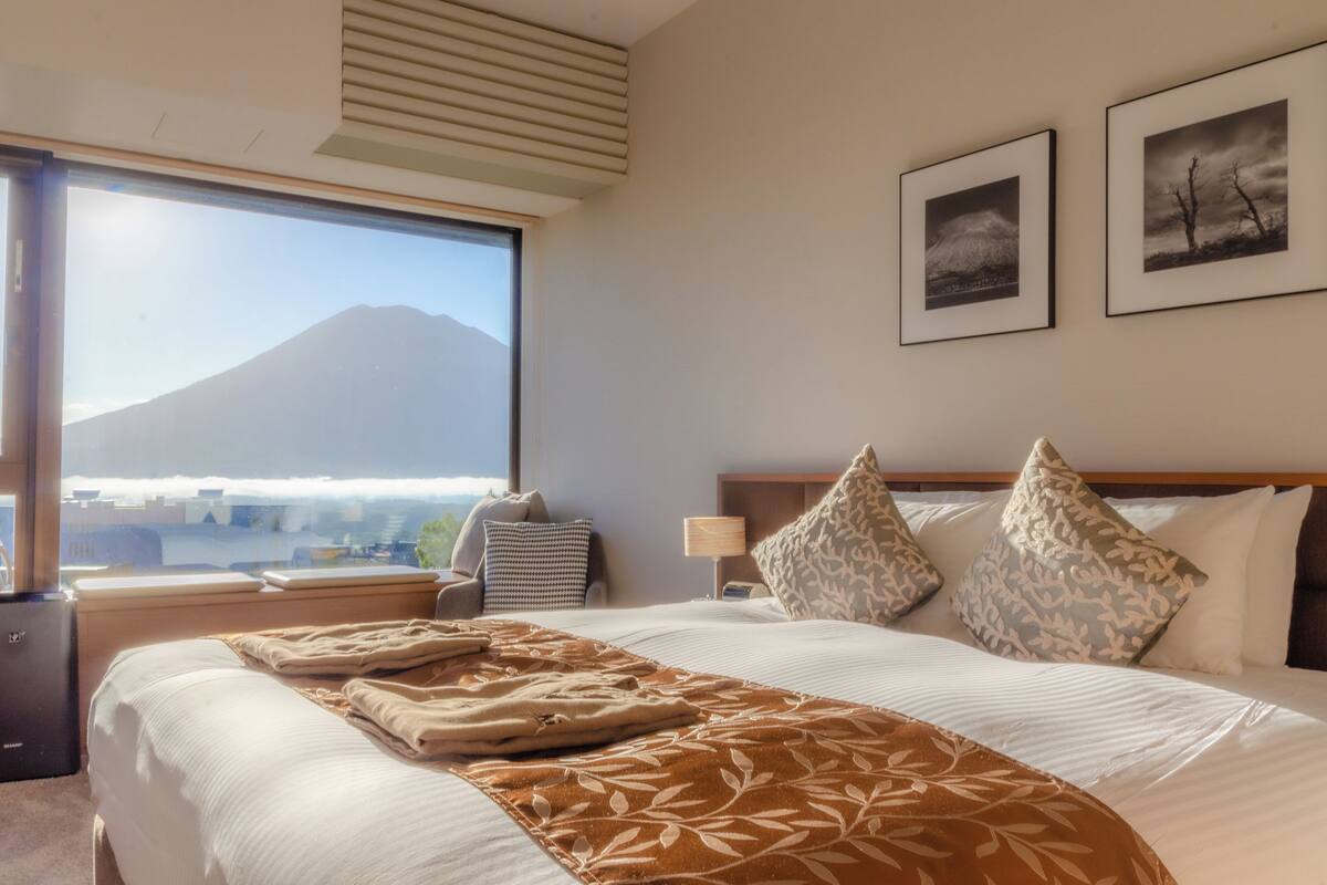 A hotel room with a large bed, a nightstand, a lamp, and a window with a view of a mountain.