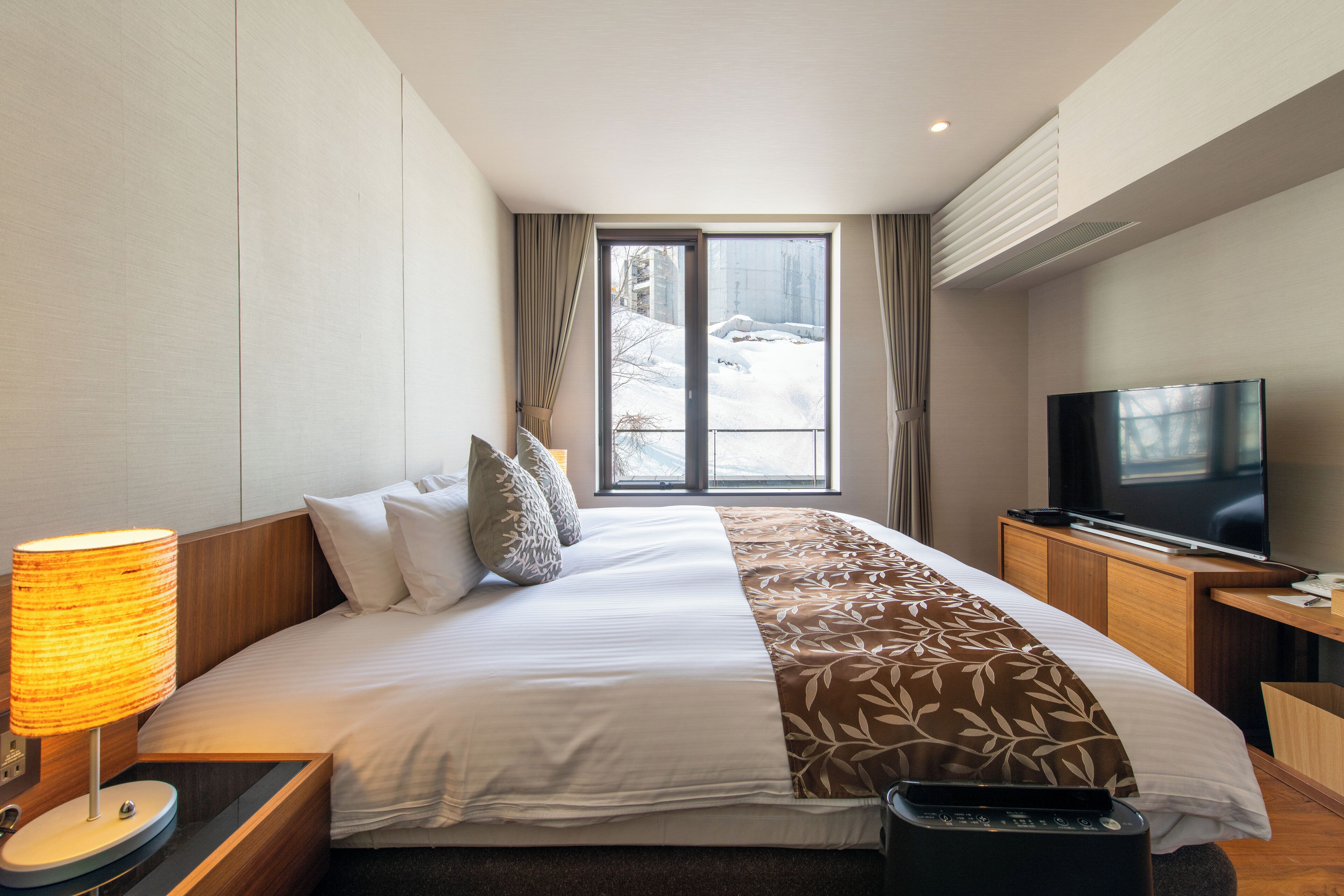 A hotel room with a large bed, a television, a lamp, and a view of the outdoors through a window.