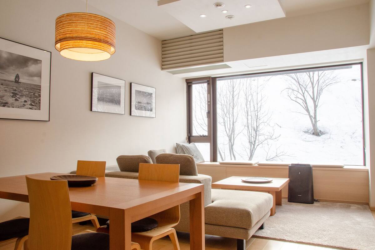 A modern living room with a wooden dining table, a sofa, and a large window with a view of trees.