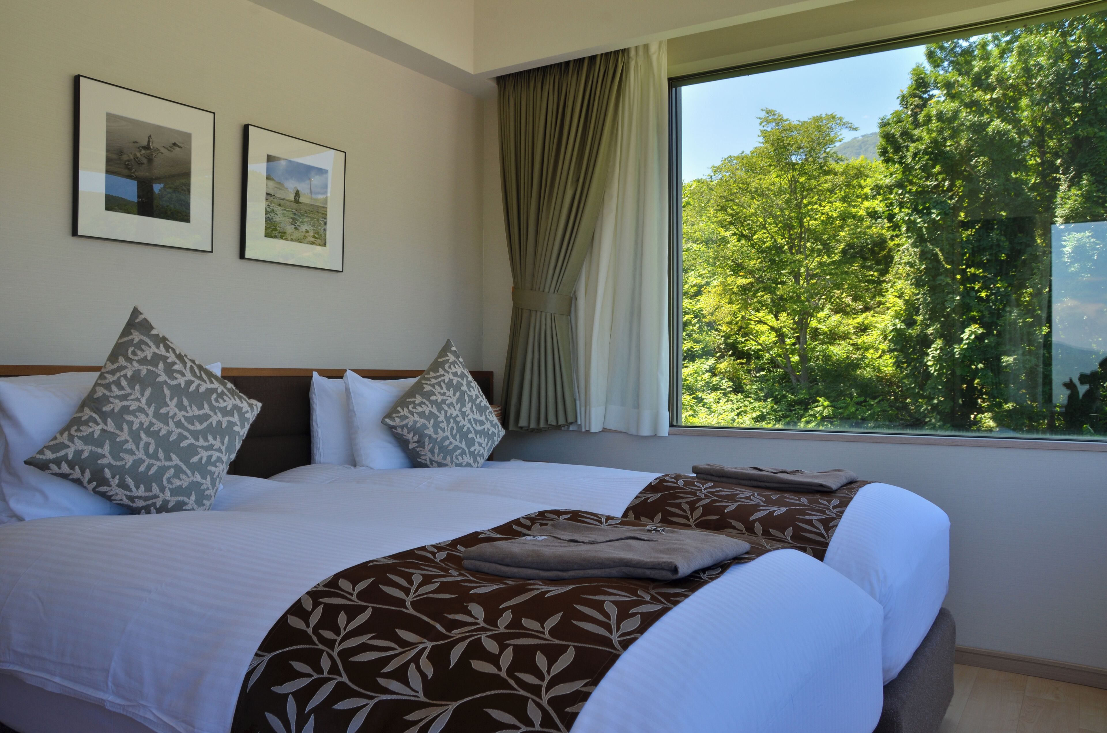 A hotel room with a large bed, two pillows, a blanket, and a view of trees through a window.