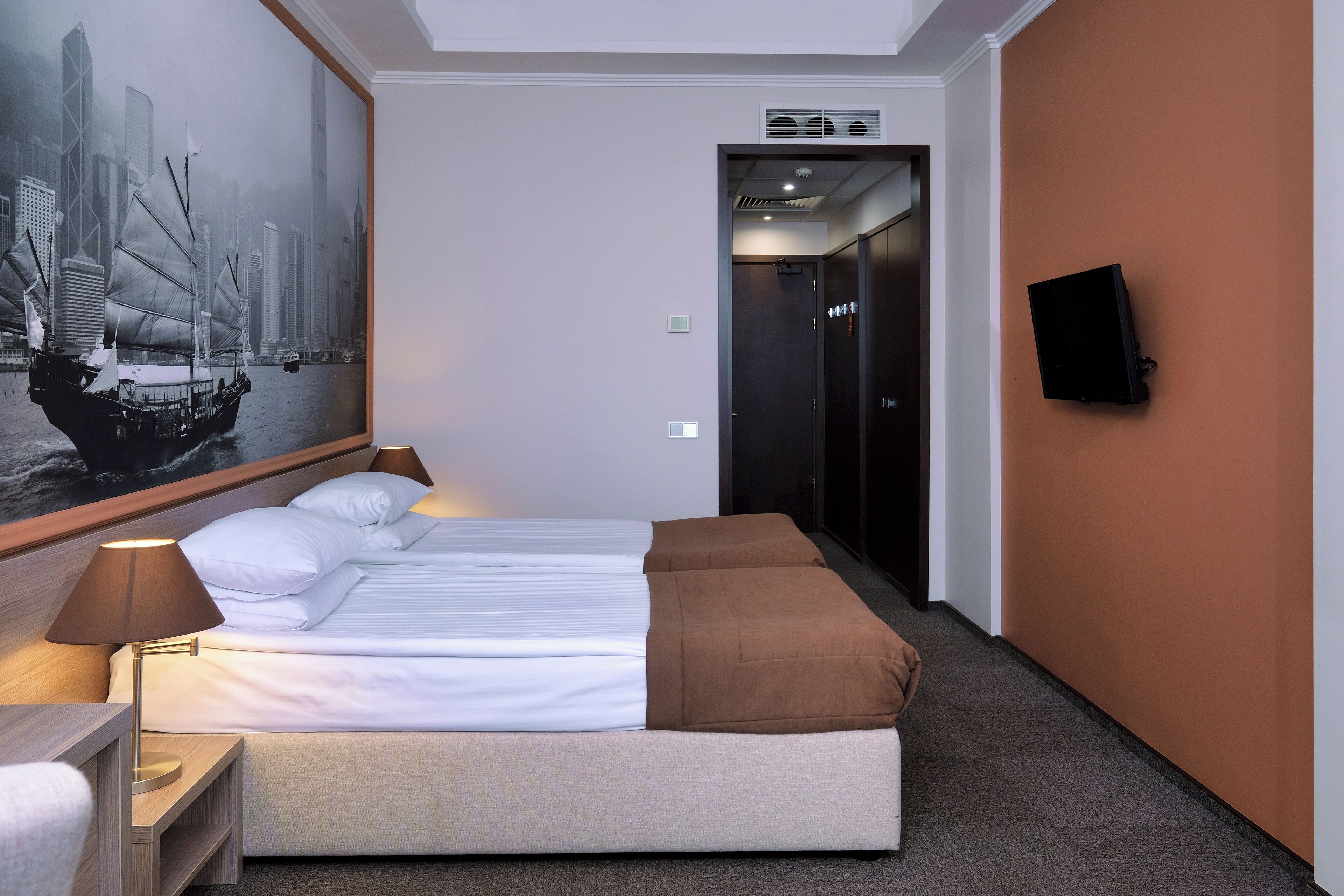 Une chambre d’hôtel avec un lit, une table de chevet, une lampe, un téléviseur fixé au mur et un grand tableau représentant un paysage urbain.