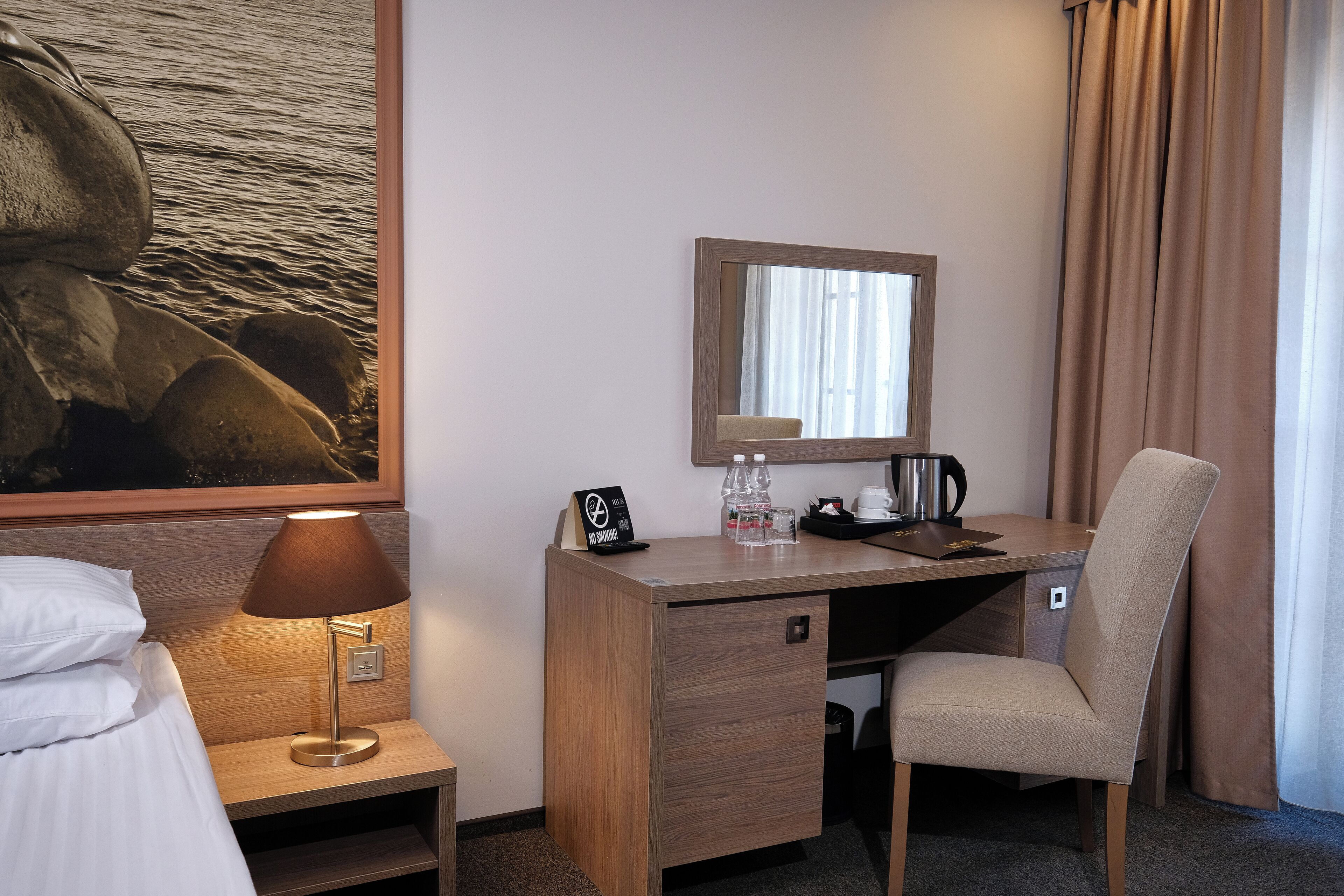 A hotel room with a bed, a desk with a lamp, a chair, a mirror, and a painting of rocks.