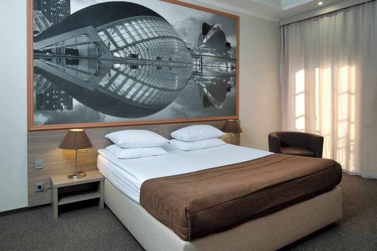 A hotel room with a large bed, two bedside tables, a lamp, a chair, and a large wall art piece.