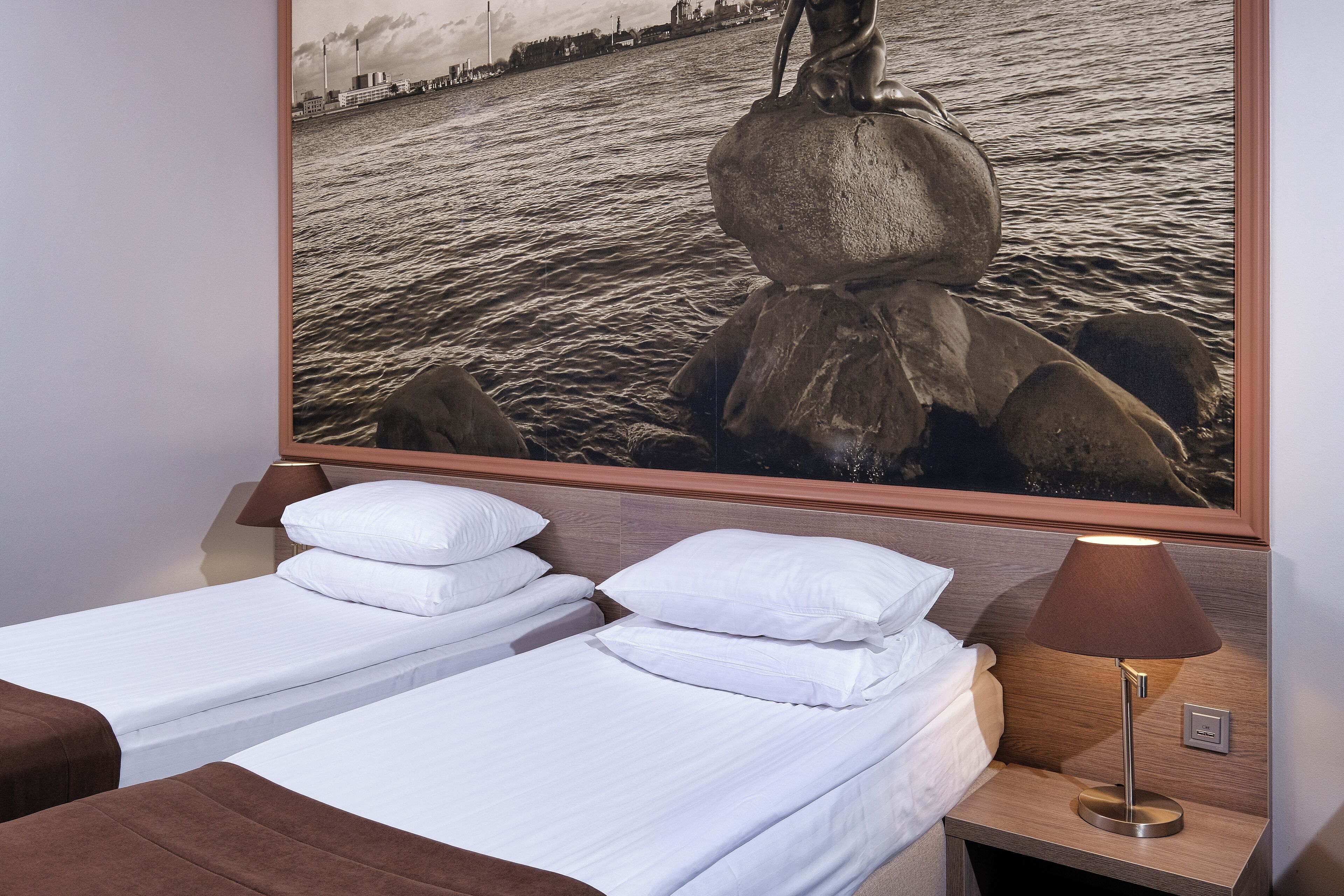 Habitación de hotel con dos camas, una mesita de noche con lámpara y un cuadro grande de una escultura junto al agua.