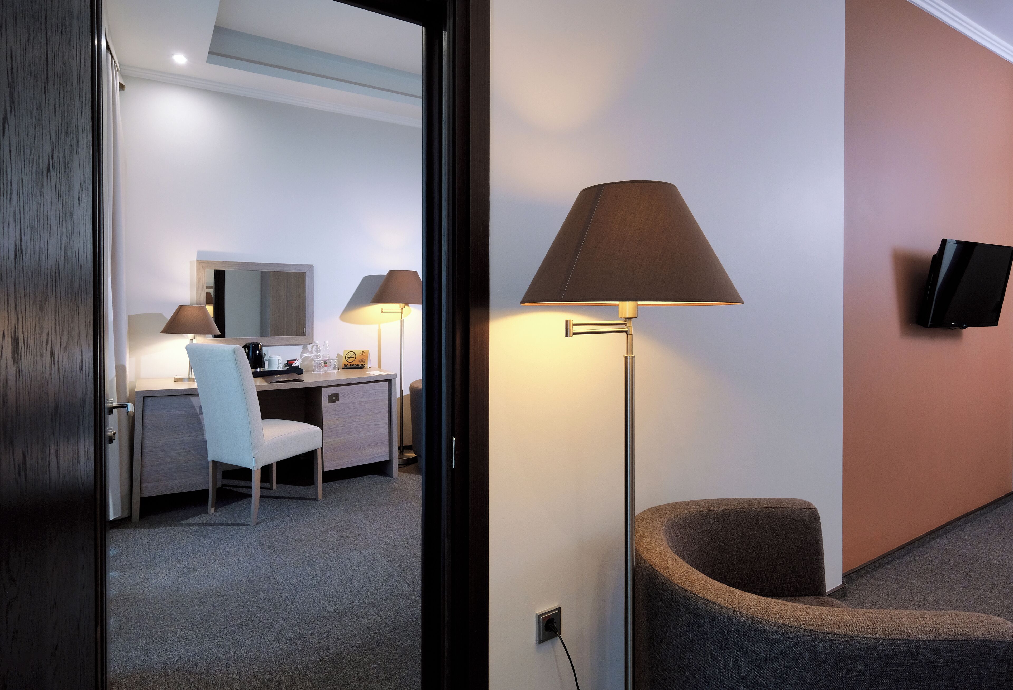 Ein modernes Hotelzimmer mit Schreibtisch, Stuhl, Nachttischlampe und einem an der Wand befestigten Flachbildfernseher.