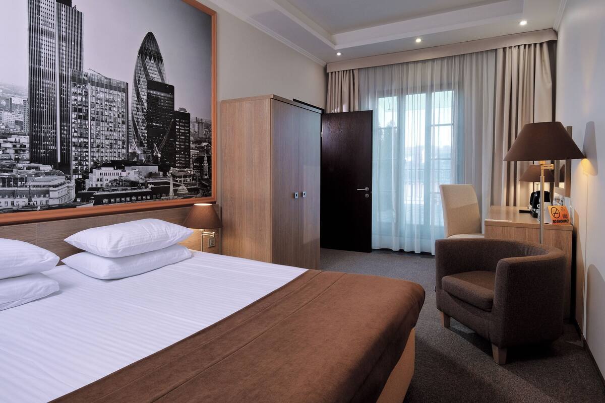 A hotel room with a large bed, a wooden wardrobe, a desk with a lamp, and a cityscape mural on the wall.