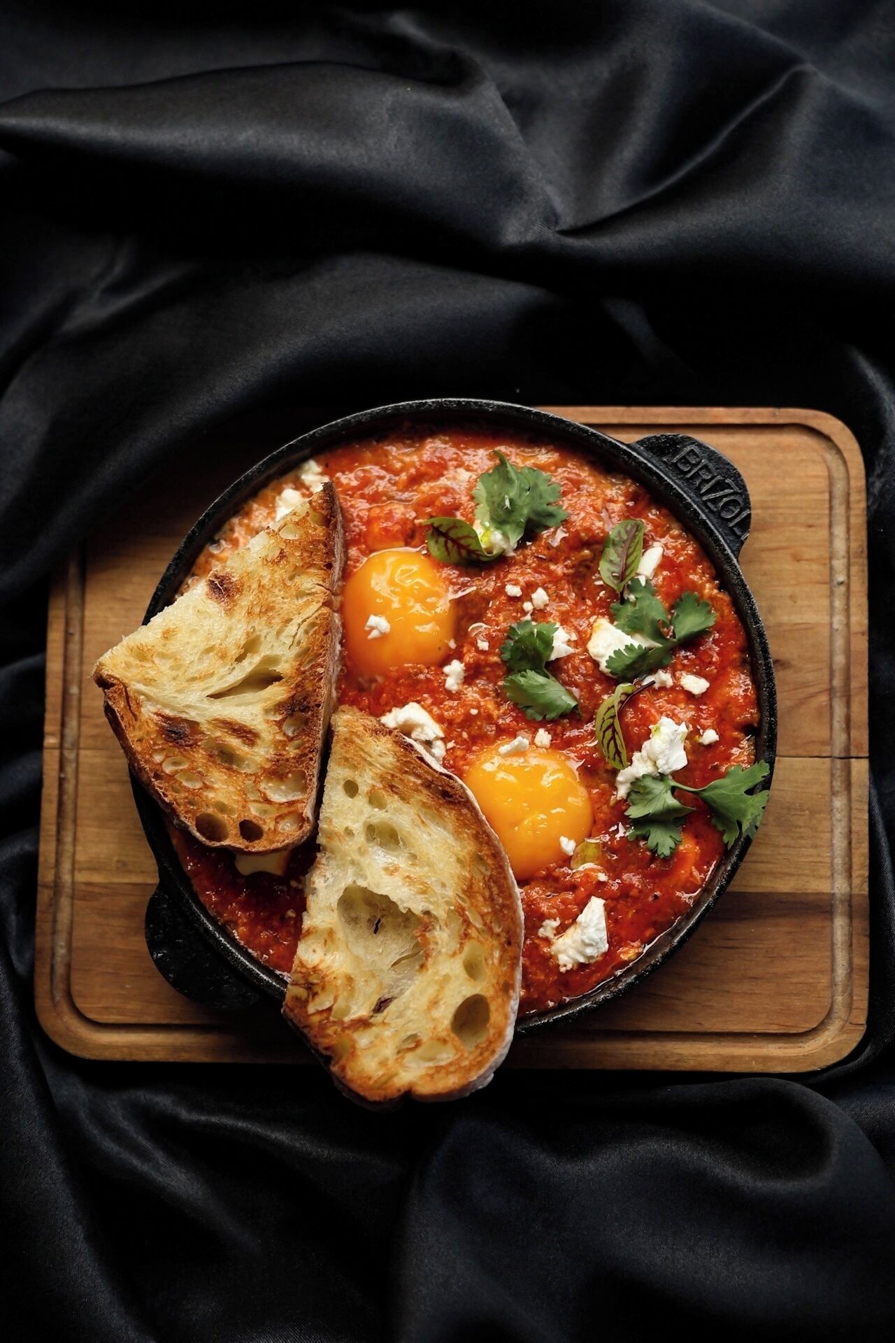 A dish of poached eggs in a red sauce, garnished with herbs, served in a black skillet on a wooden board.
