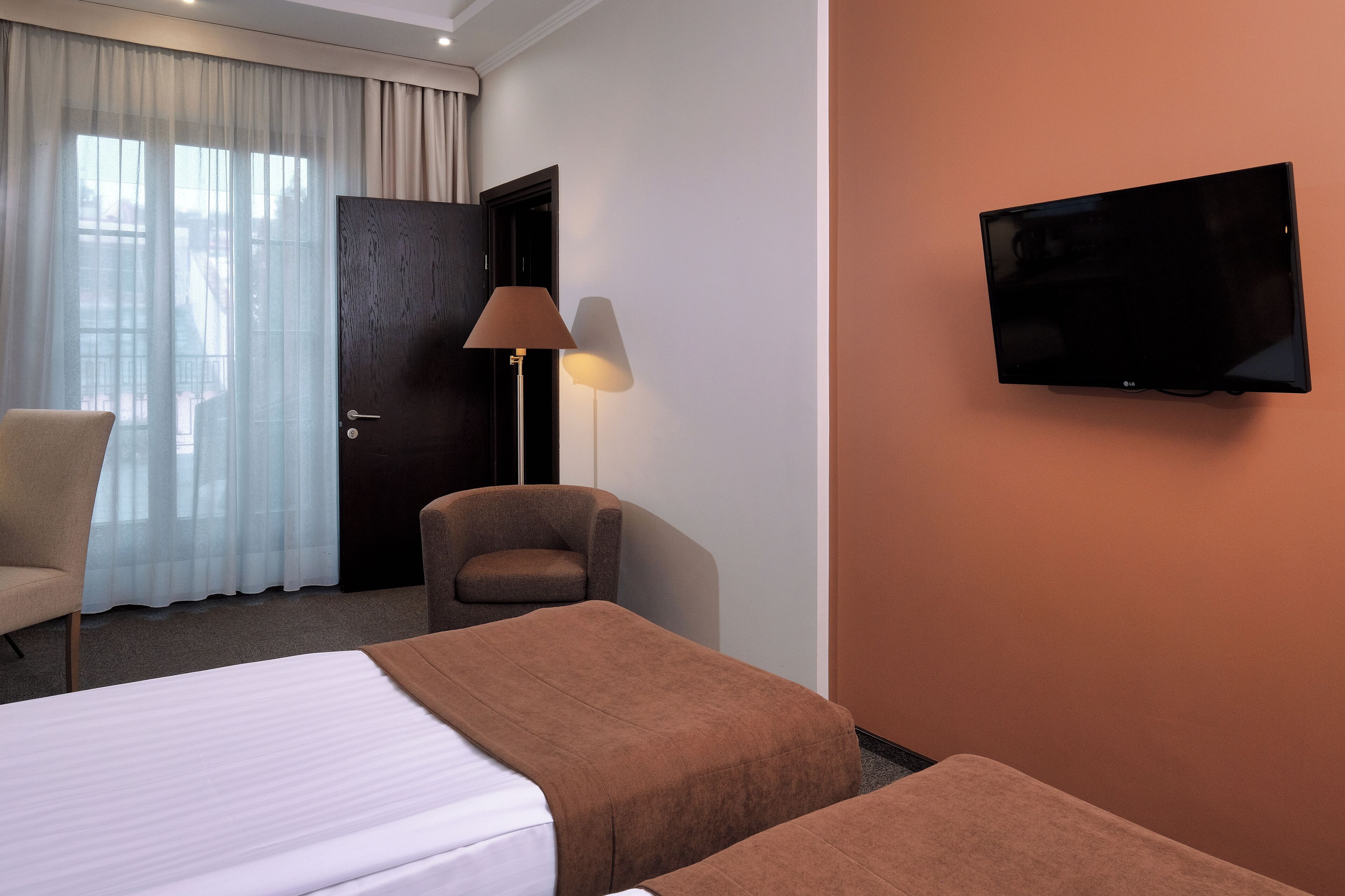 A hotel room with a bed, two bedside tables, a chair, a desk, a lamp, and a TV mounted on the wall.