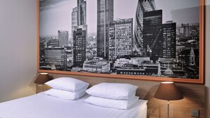 A hotel room with a bed, two bedside lamps, and a large cityscape poster on the wall.