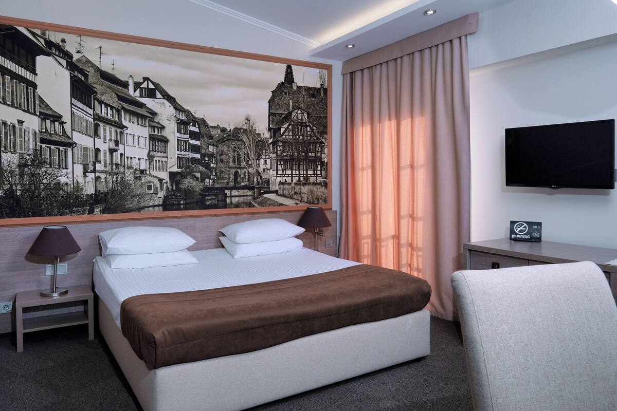 A hotel room with a large bed, a desk with a flat-screen TV, a painting of a European town, and a view of the city through the window.