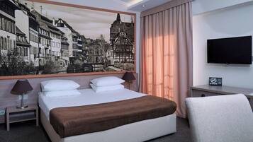 Quarto de hotel com uma cama grande, uma escrivaninha com televisão de ecrã plano, um quadro de uma cidade europeia e vista para a cidade pela janela.