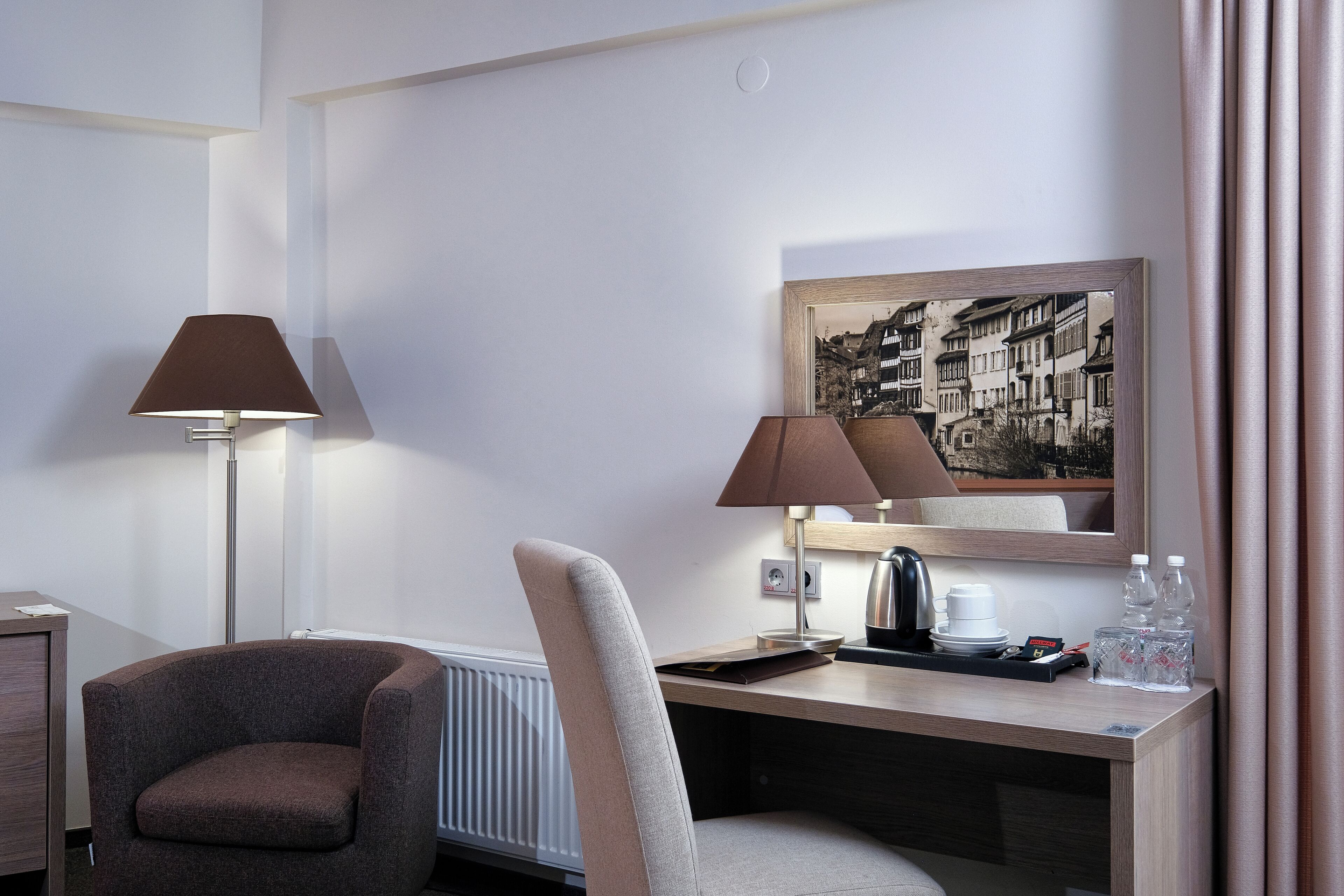 A hotel room with a desk, two lamps, a chair, a framed picture, and a radiator.