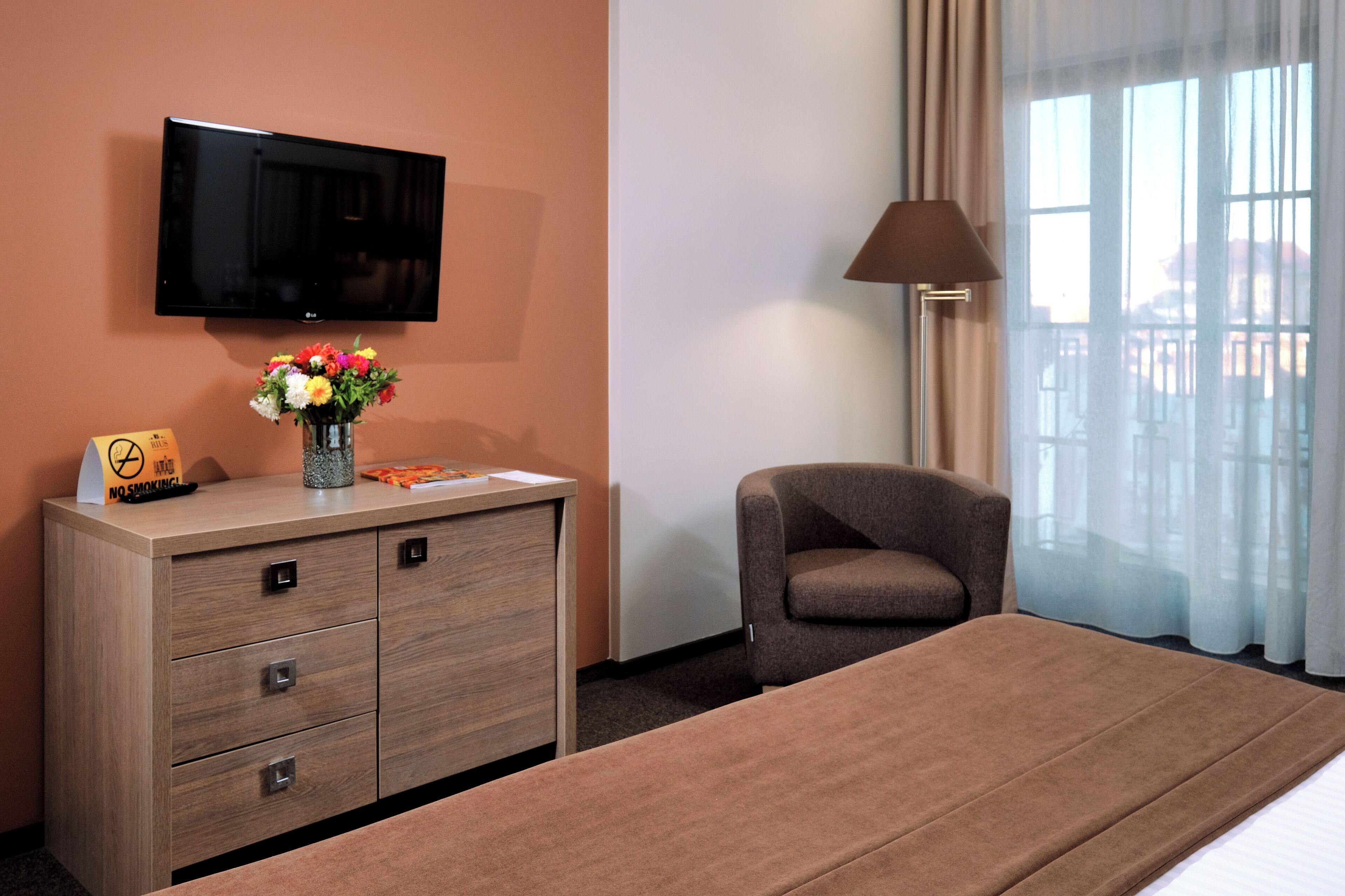 A hotel room with a bed, a TV mounted on the wall, a desk with a vase of flowers, a chair, and a lamp.
