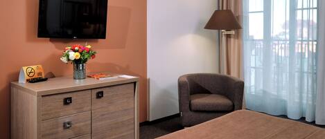 A hotel room with a bed, a TV mounted on the wall, a desk with a vase of flowers, a chair, and a lamp.