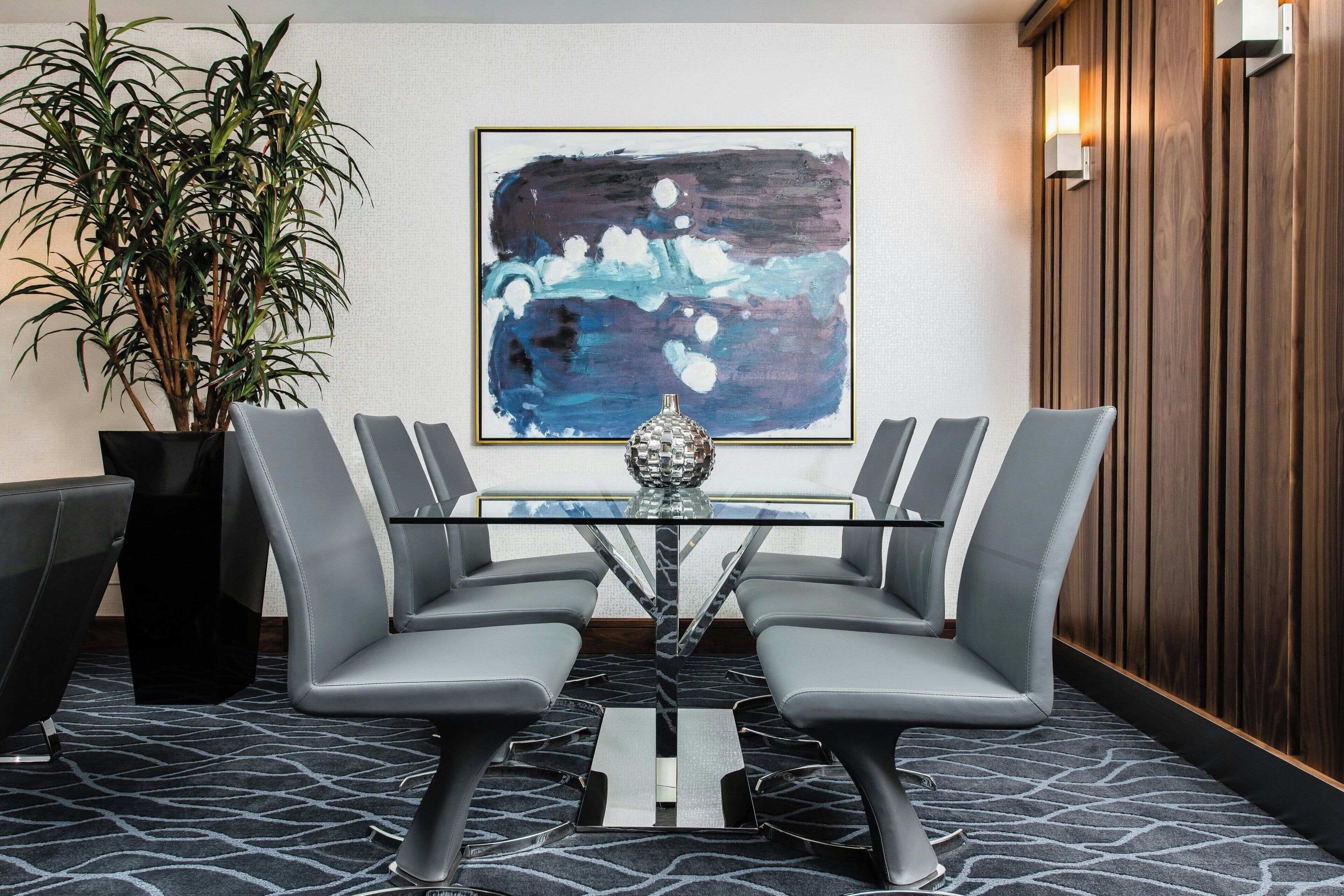 A modern conference room with a glass table, grey chairs, a large abstract painting, a potted plant, and a wooden panel wall.