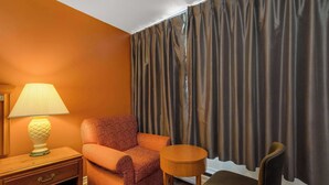 A room with an orange wall, a patterned armchair, a wooden nightstand with a lamp, a small round table, and a bench. - Knights Inn Burlington, ON (Burlington)