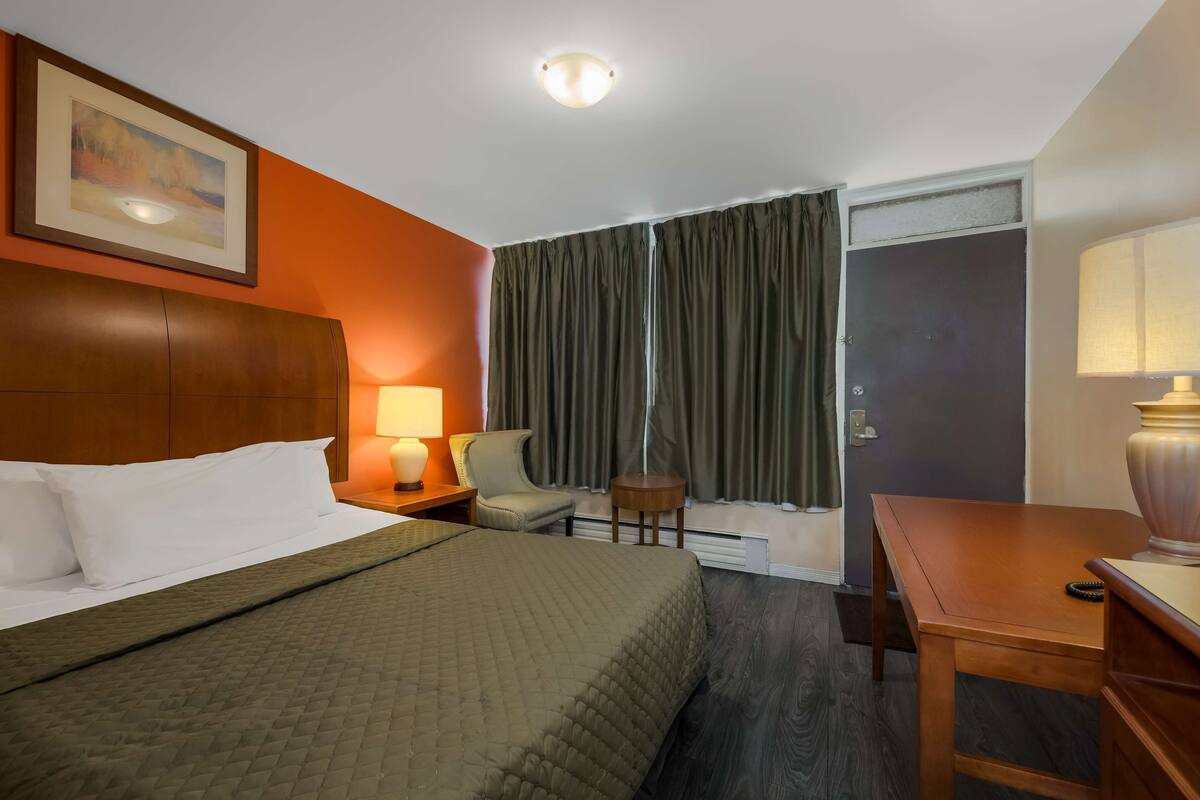A hotel room with a bed, a desk, a chair, a lamp, and a painting on the wall.