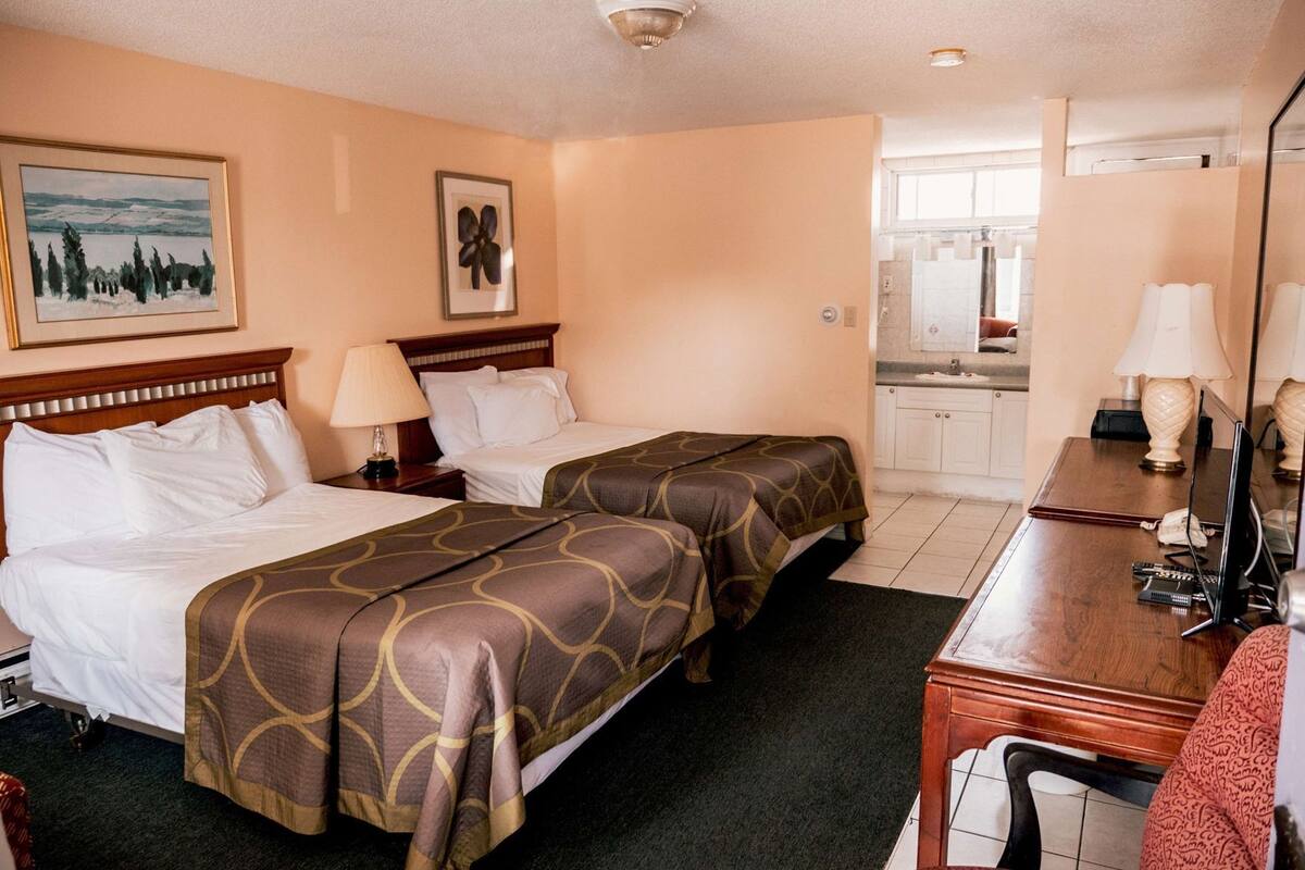 A hotel room with two beds, a desk, a chair, a lamp, a painting, and a bathroom visible through an open door.