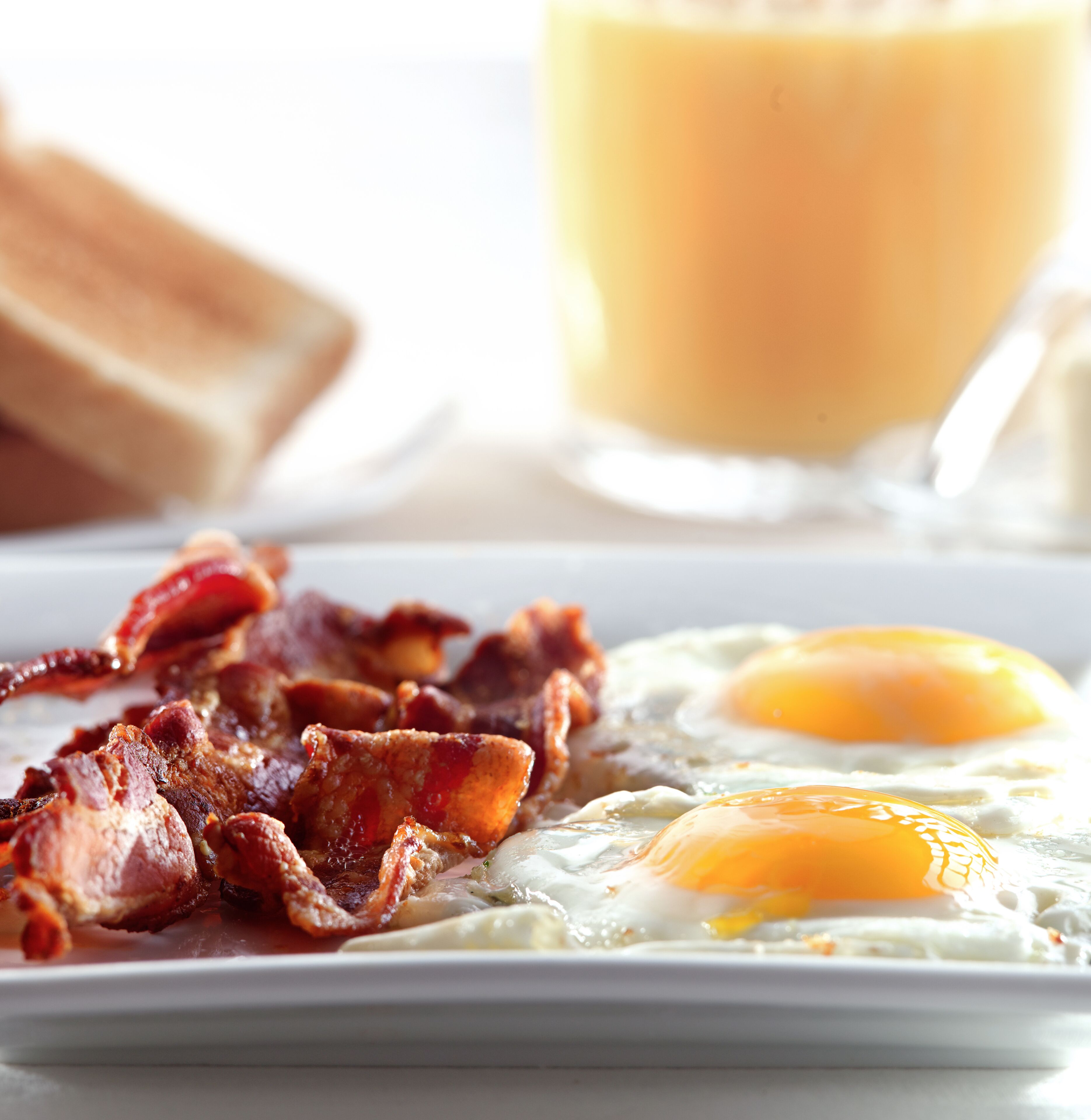 Une assiette de petit-déjeuner avec du bacon et des œufs, un verre de jus d’orange et une planche à découper en bois en arrière-plan.