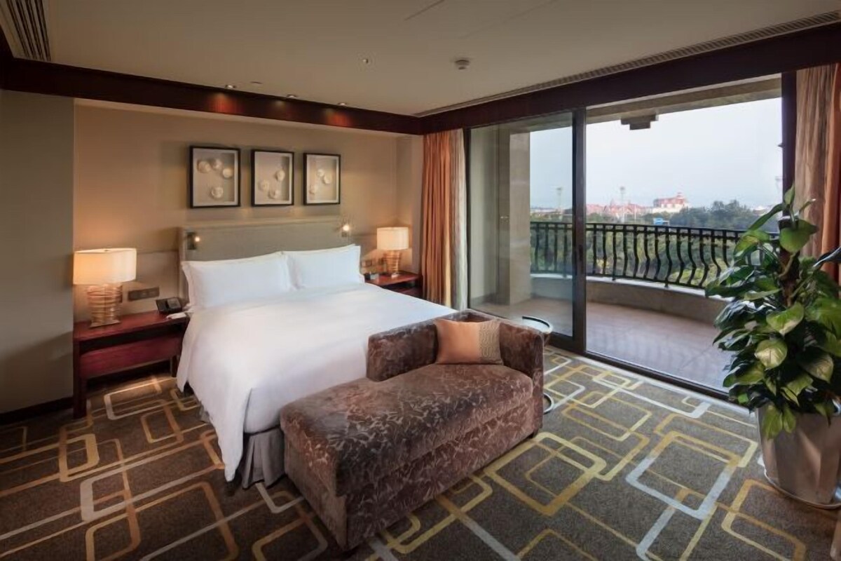 A hotel room with a large bed, a sofa, a balcony with a view, and a potted plant.